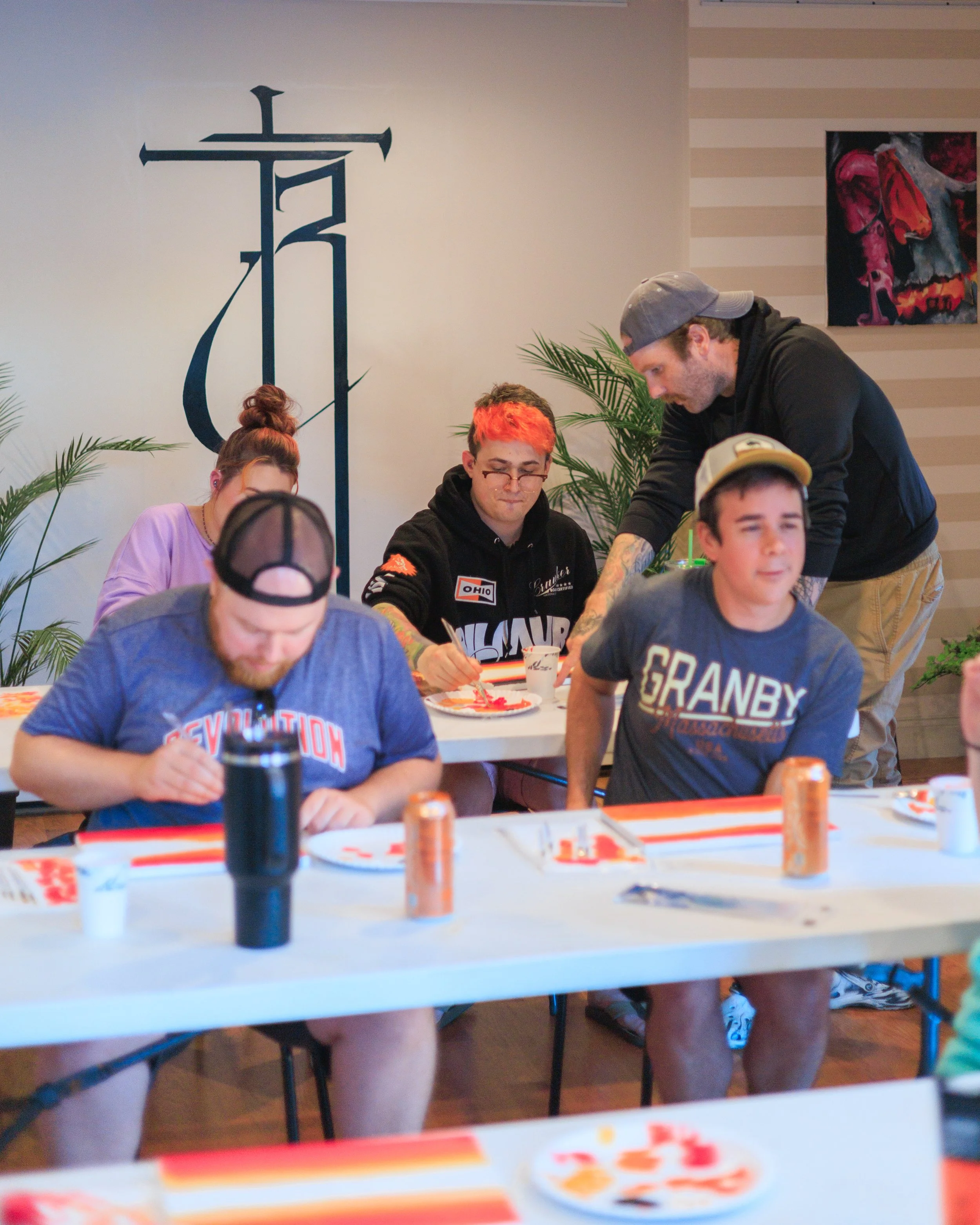 Group of young people sitting at a table during a meal, with one person standing helping another. There are plates, drinks, and a can of soda on the table, with a large cross symbol on the wall behind them.