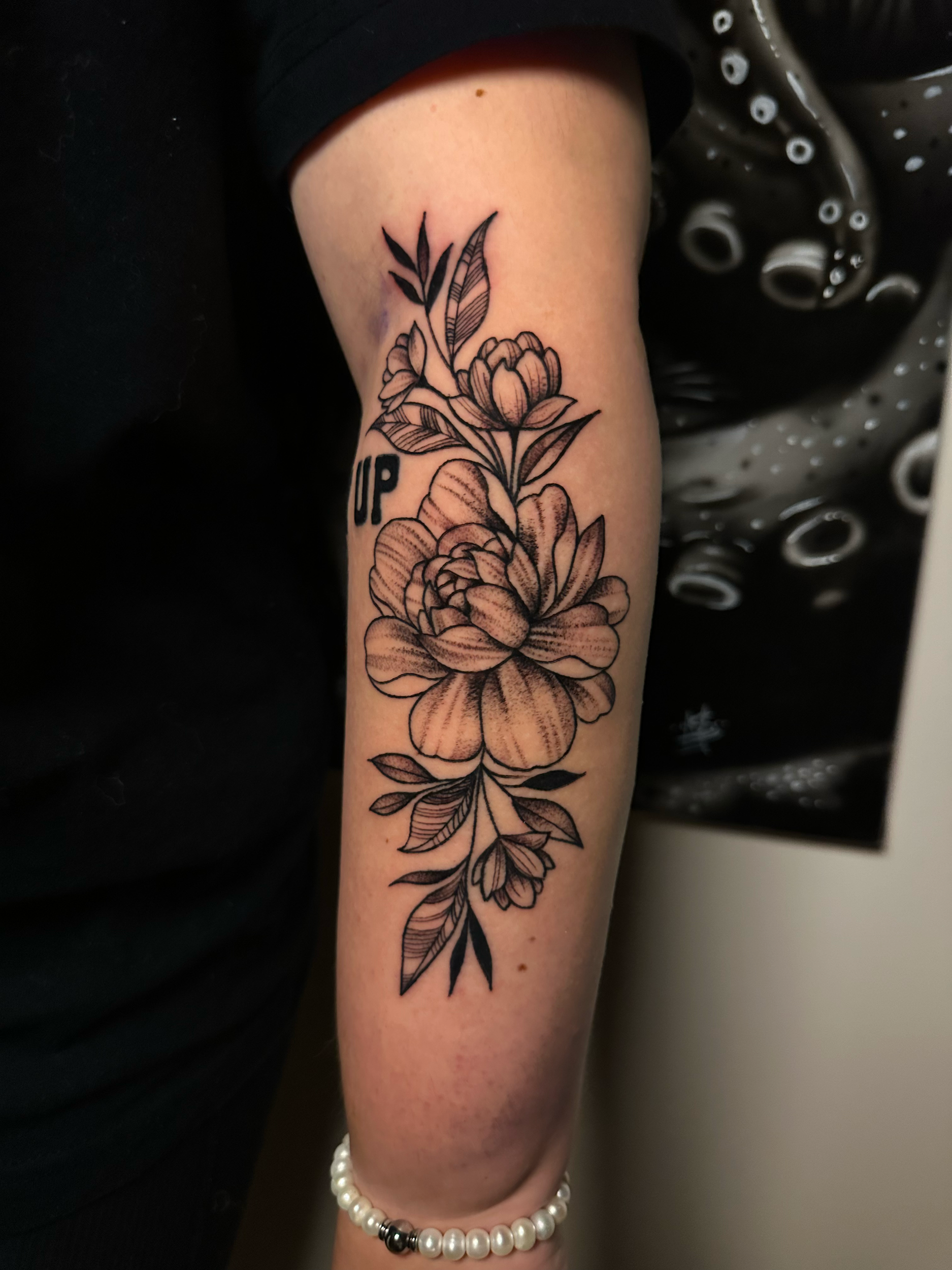 Floral tattoo on forearm featuring roses and leaves in black ink, person wearing a pearl bracelet.