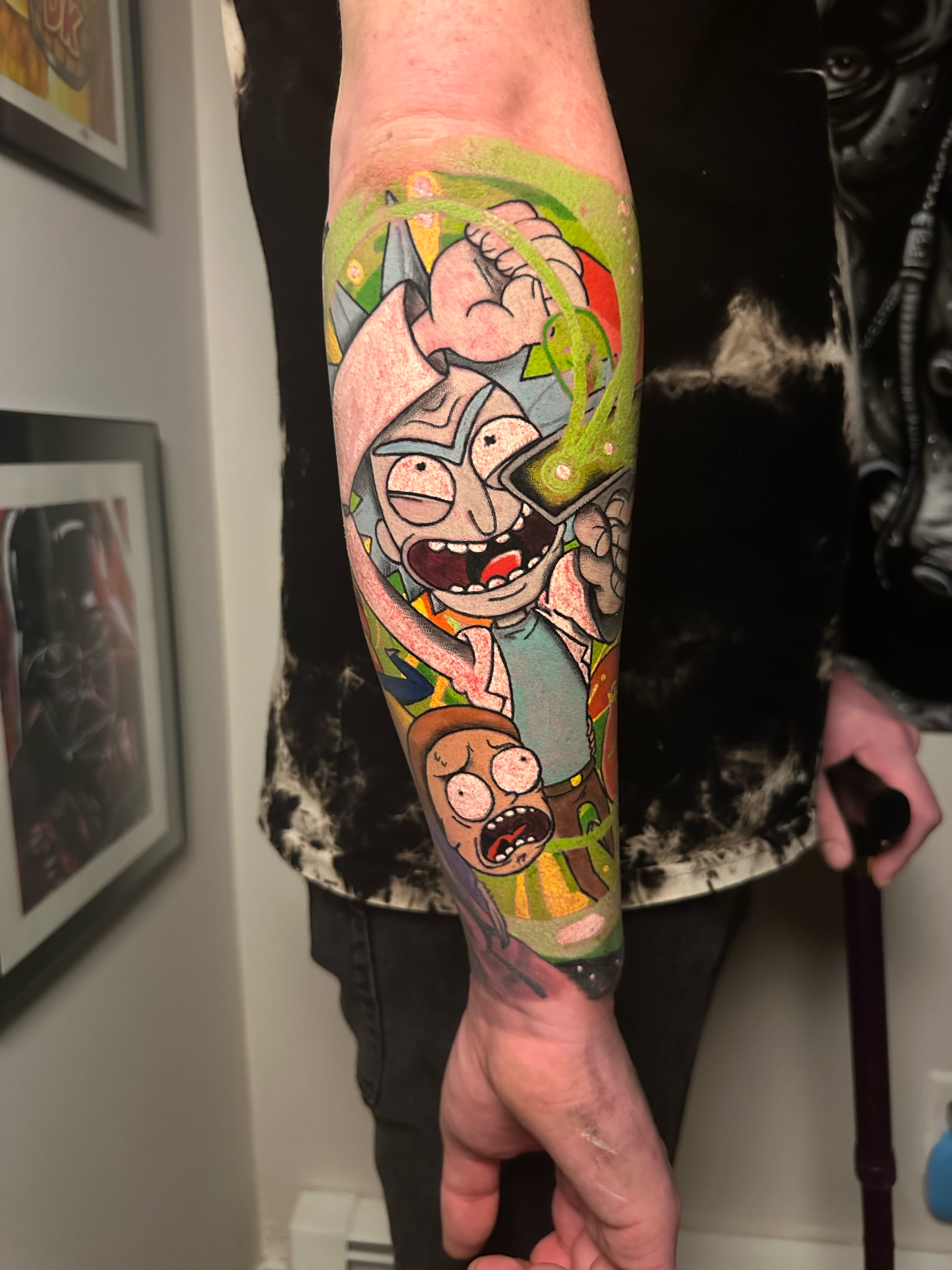 Colorful cartoon-style forearm tattoo featuring two animated characters with exaggerated facial expressions. The artwork includes a background with bright colors and abstract designs.