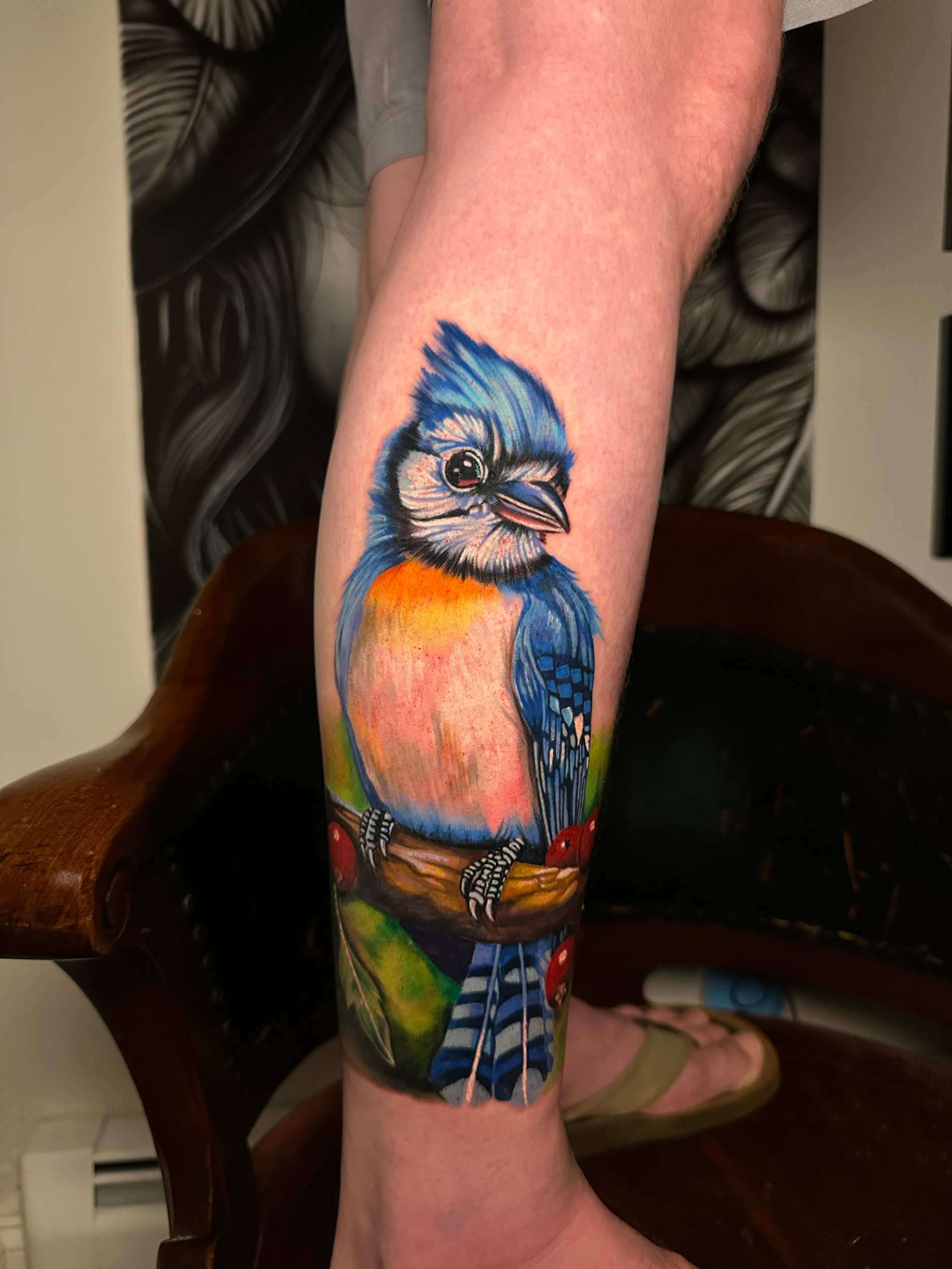 Colorful tattoo of a bird with bright blue, orange, and black feathers sitting on a branch with red berries.