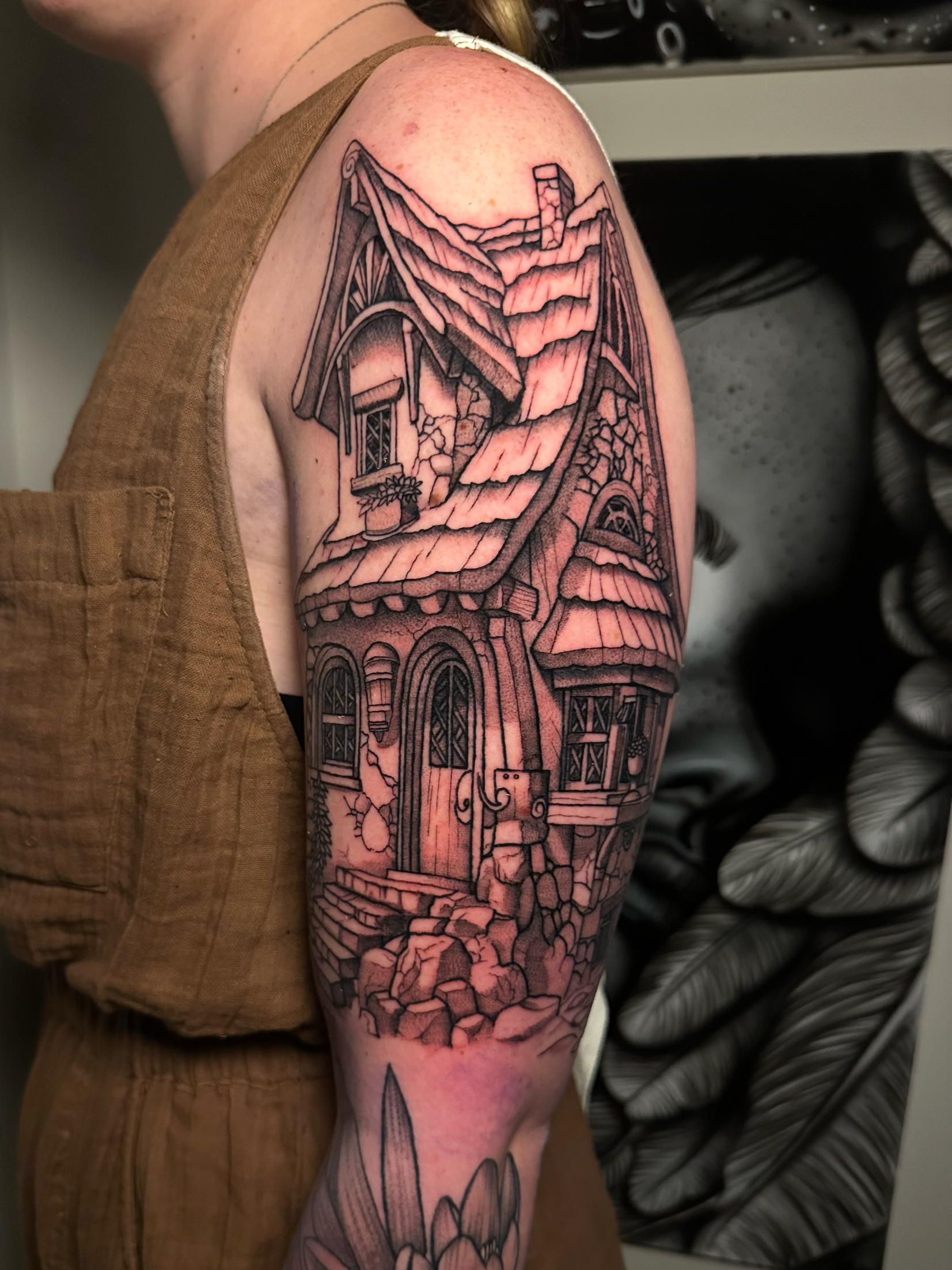 Black and gray tattoo of a detailed house with a roof, chimney, windows, and a front door on a person's upper arm.