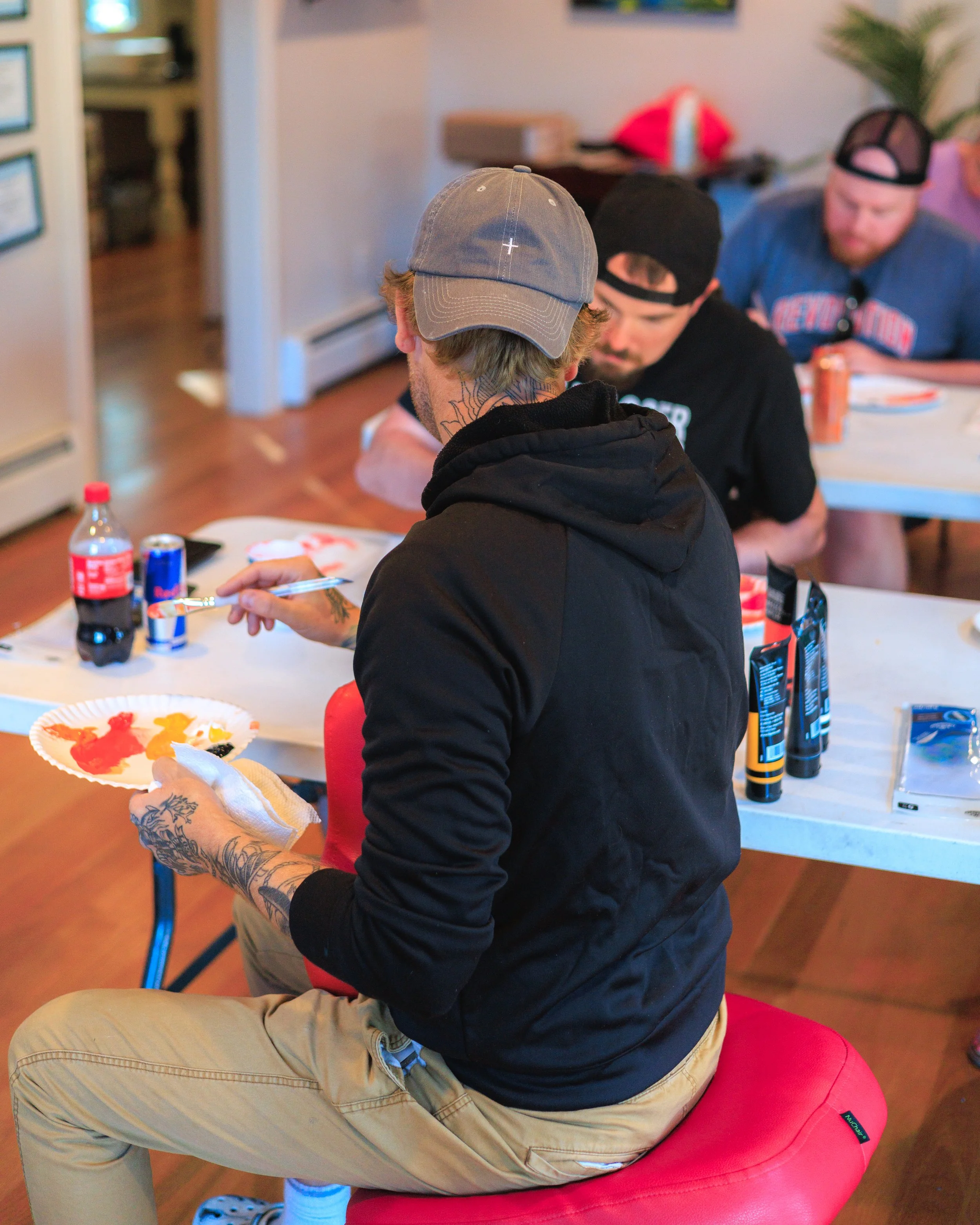 A man with tattoos on his hands and neck sits on a red cushioned stool, wearing a gray cap, black hoodie, and beige pants, holding a paper plate with red and yellow paint and a napkin. He is painting or decorating the plate. In the background, others