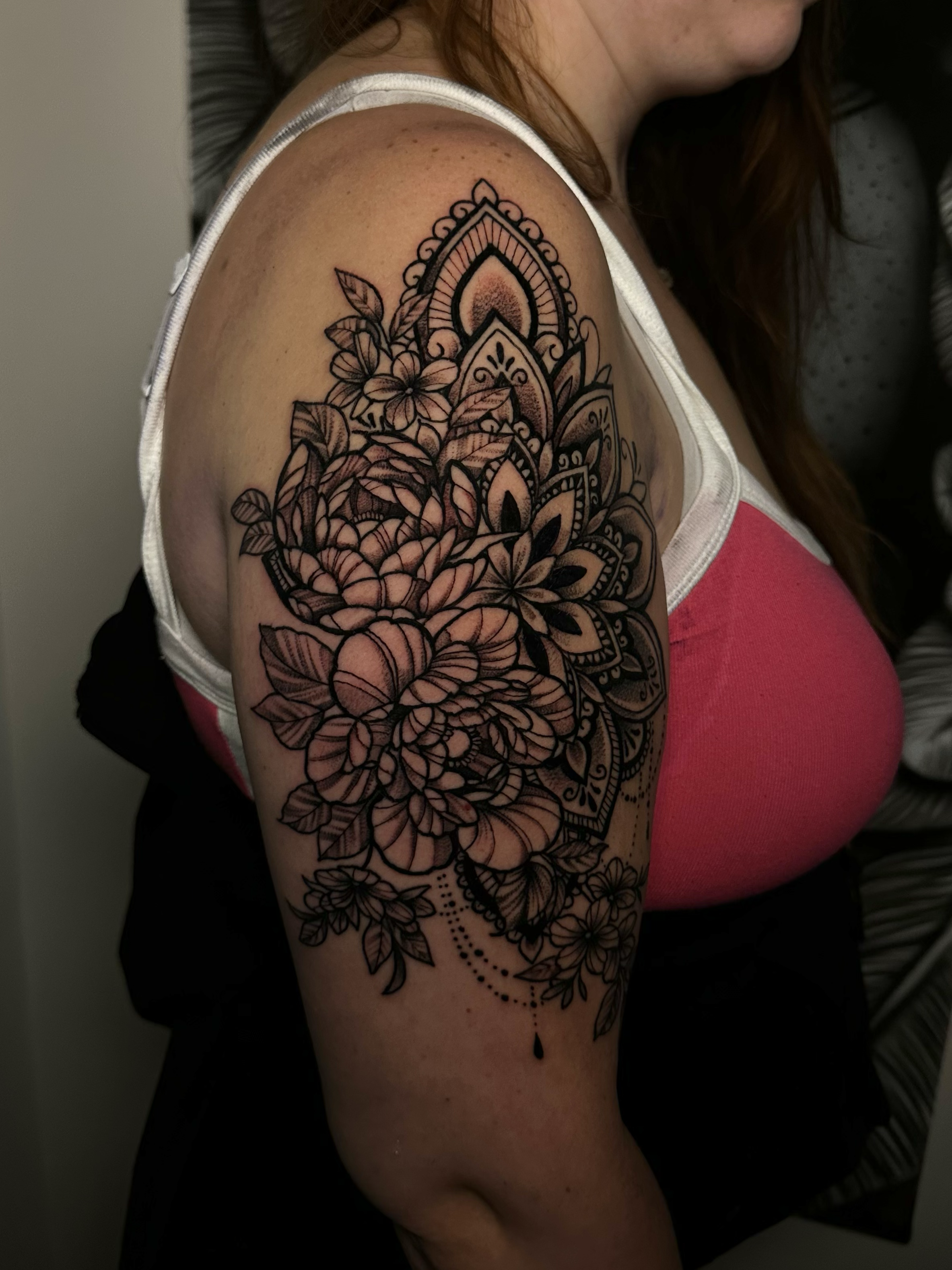 A woman with long reddish hair showing a large, intricate black and gray floral tattoo on her upper arm and shoulder.