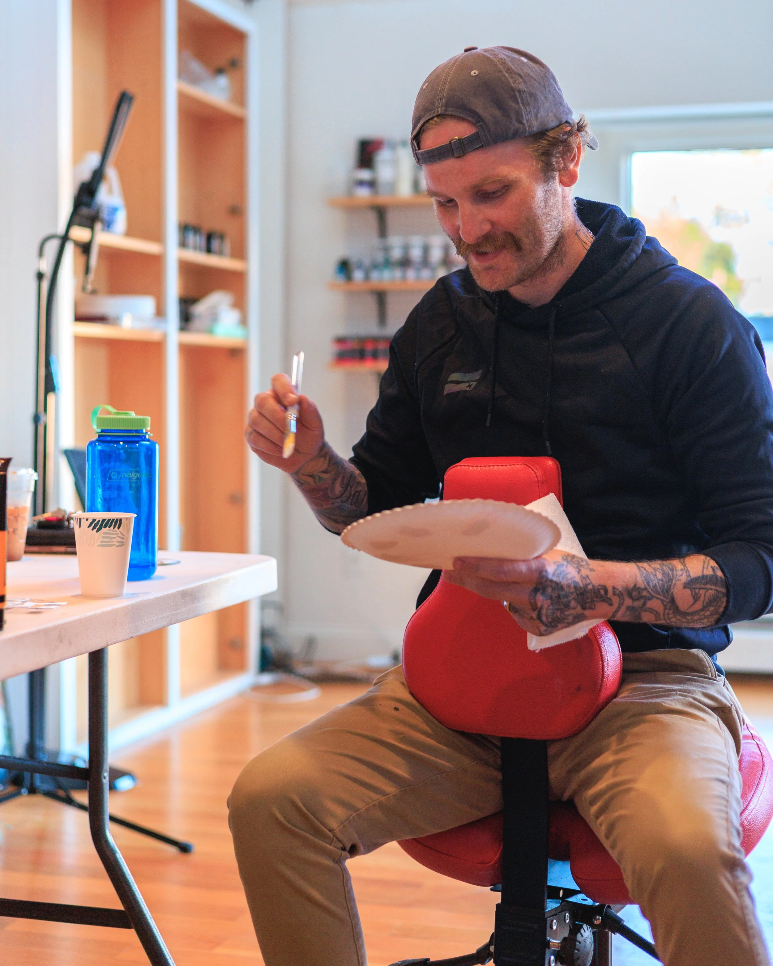 A man with tattoos on his arms, wearing a backward gray cap and a black hoodie, is sitting on a red dental chair. He is smiling and looking at a paper plate while holding a paintbrush. There is a table with various items, including a blue water bottl