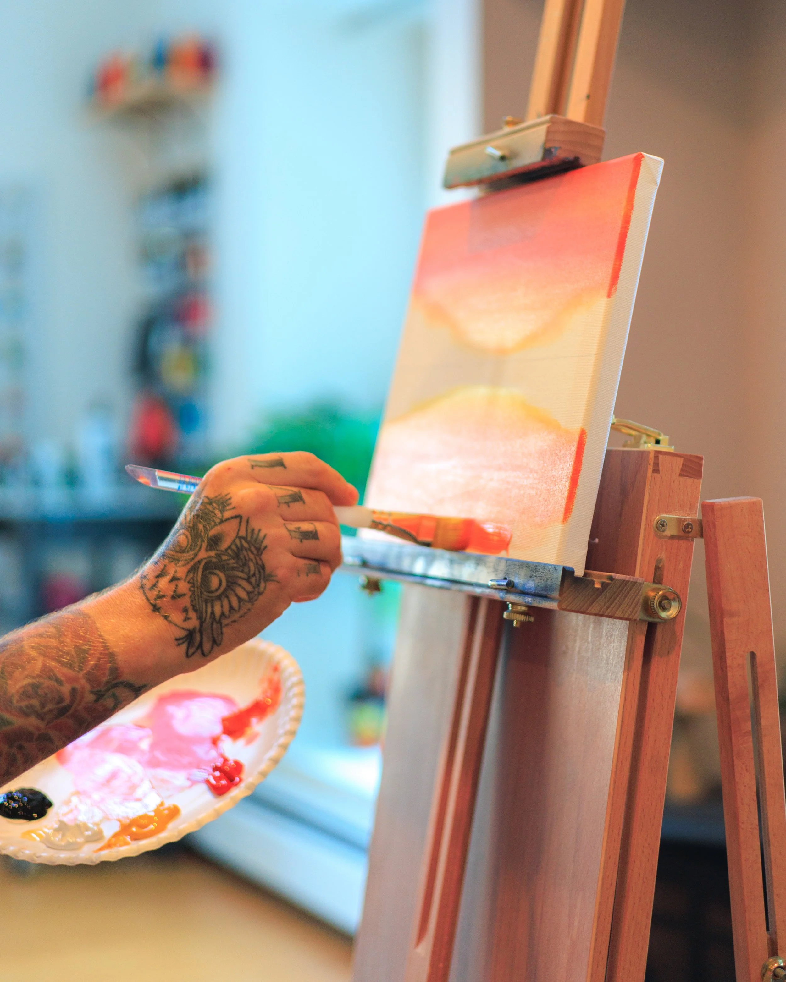 Tattooed hand holding a paintbrush, painting a sunset landscape on a canvas atop an easel in an art studio.