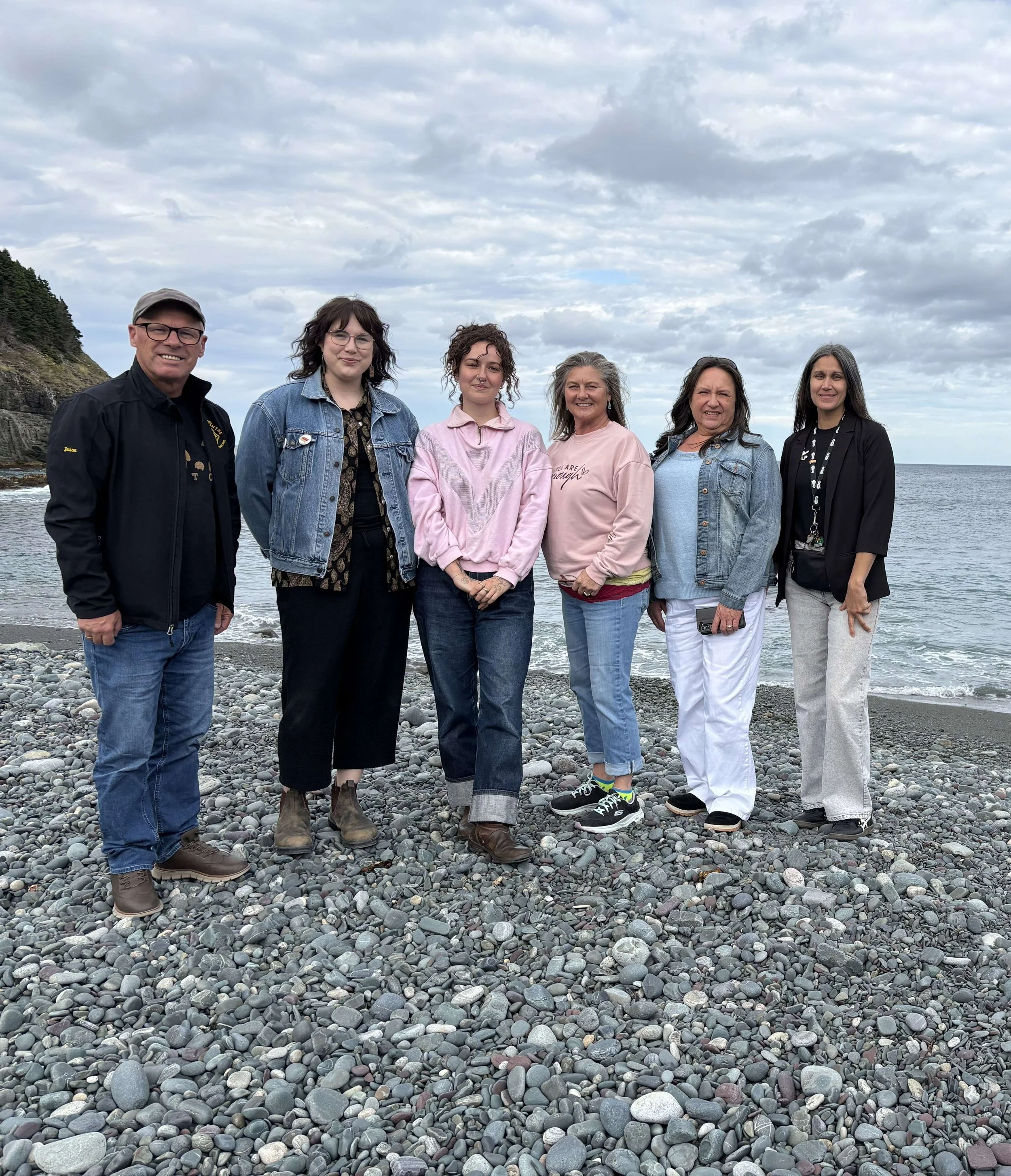 Group of six people standing on a rocky beach near the ocean under a cloudy sky.
