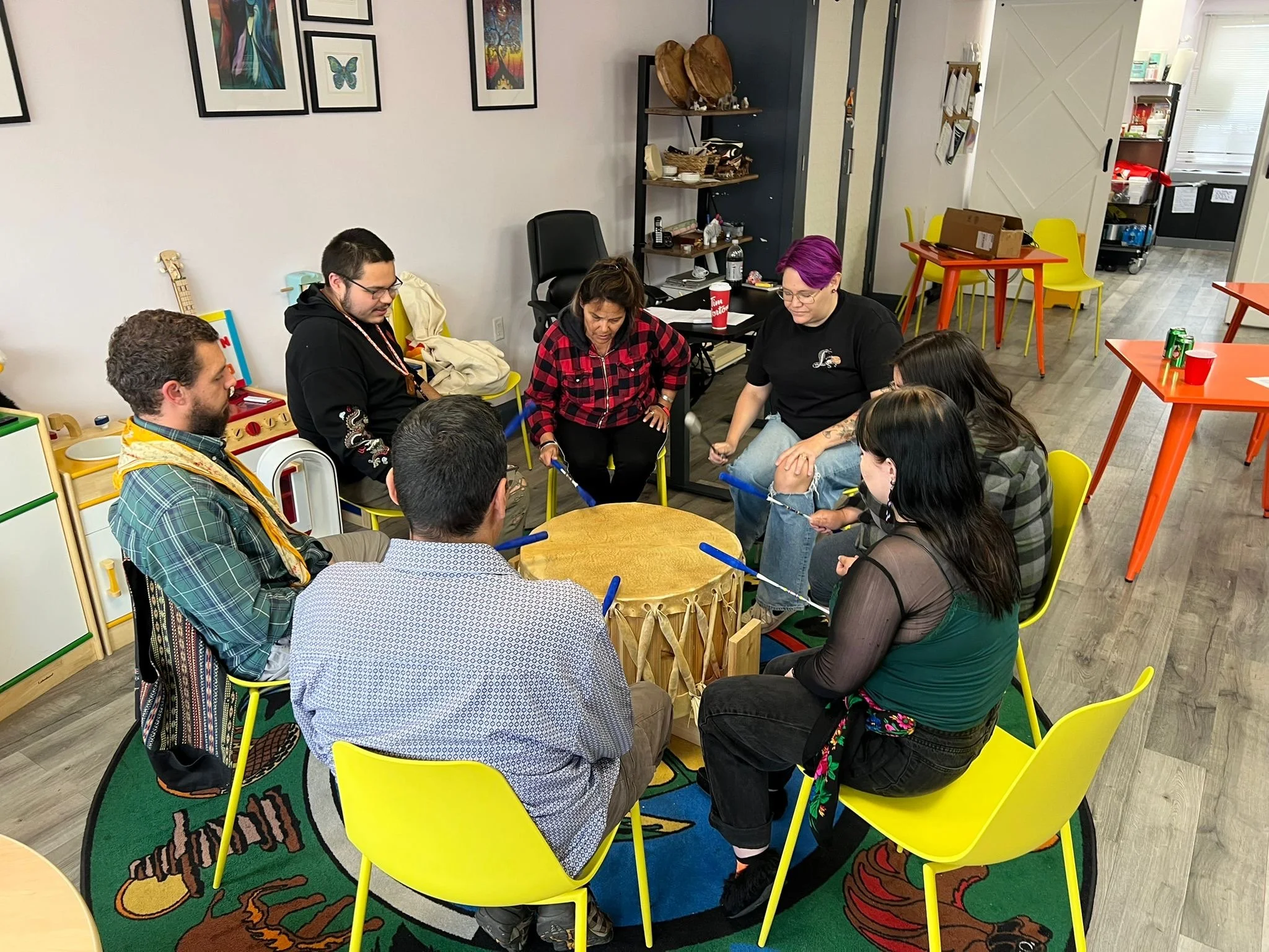 Group of people sitting in a circle around a drum in a colorful room, engaging in a group activity.