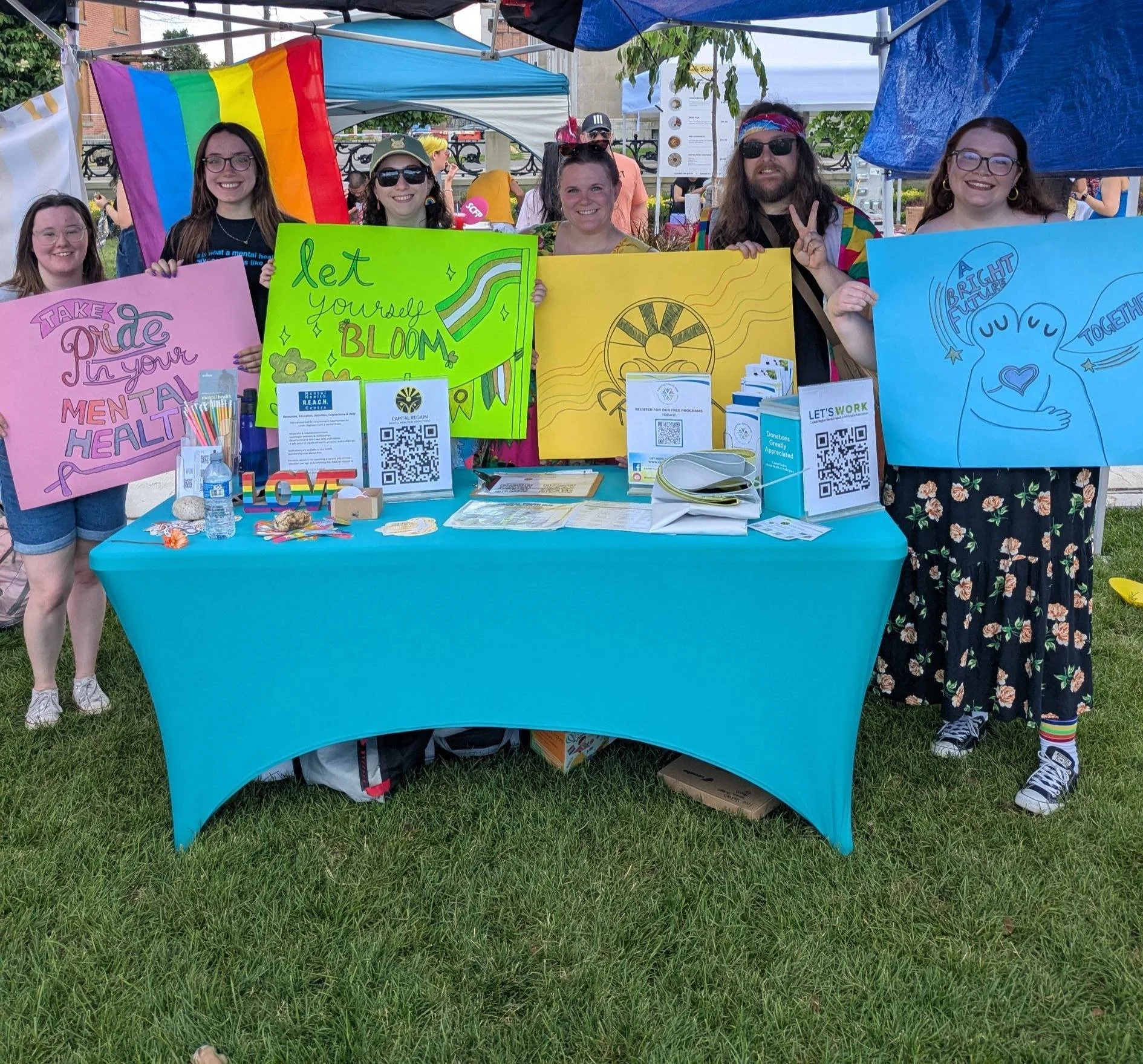 Group of people standing behind a blue table at an outdoor event, holding colorful signs with messages about mental health, pride, and community. The table has informational materials and QR codes, with a rainbow pride flag visible.