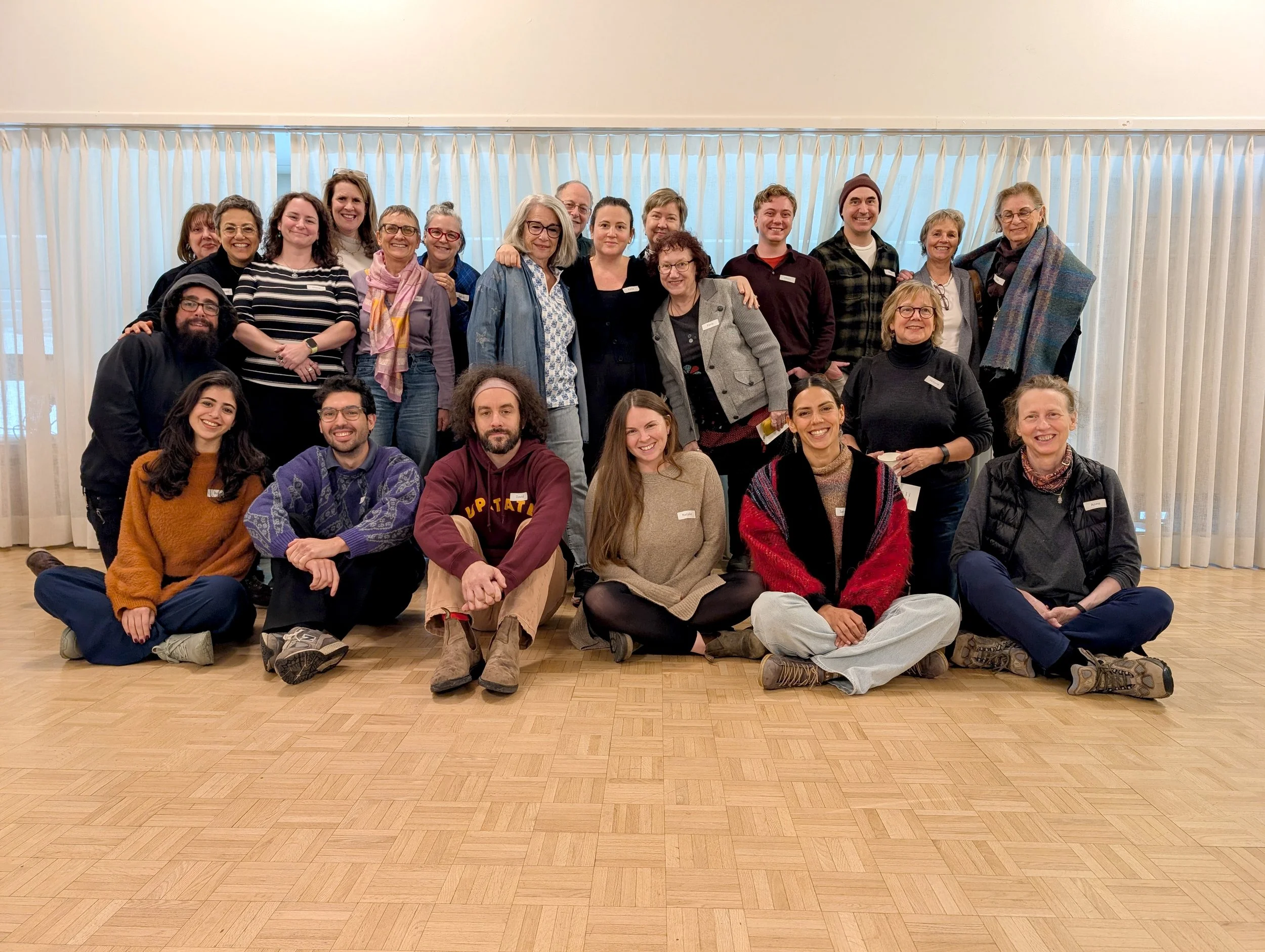 Group of 21 people posing indoors in front of white curtains, some sitting on the floor and others standing behind, smiling for the camera.