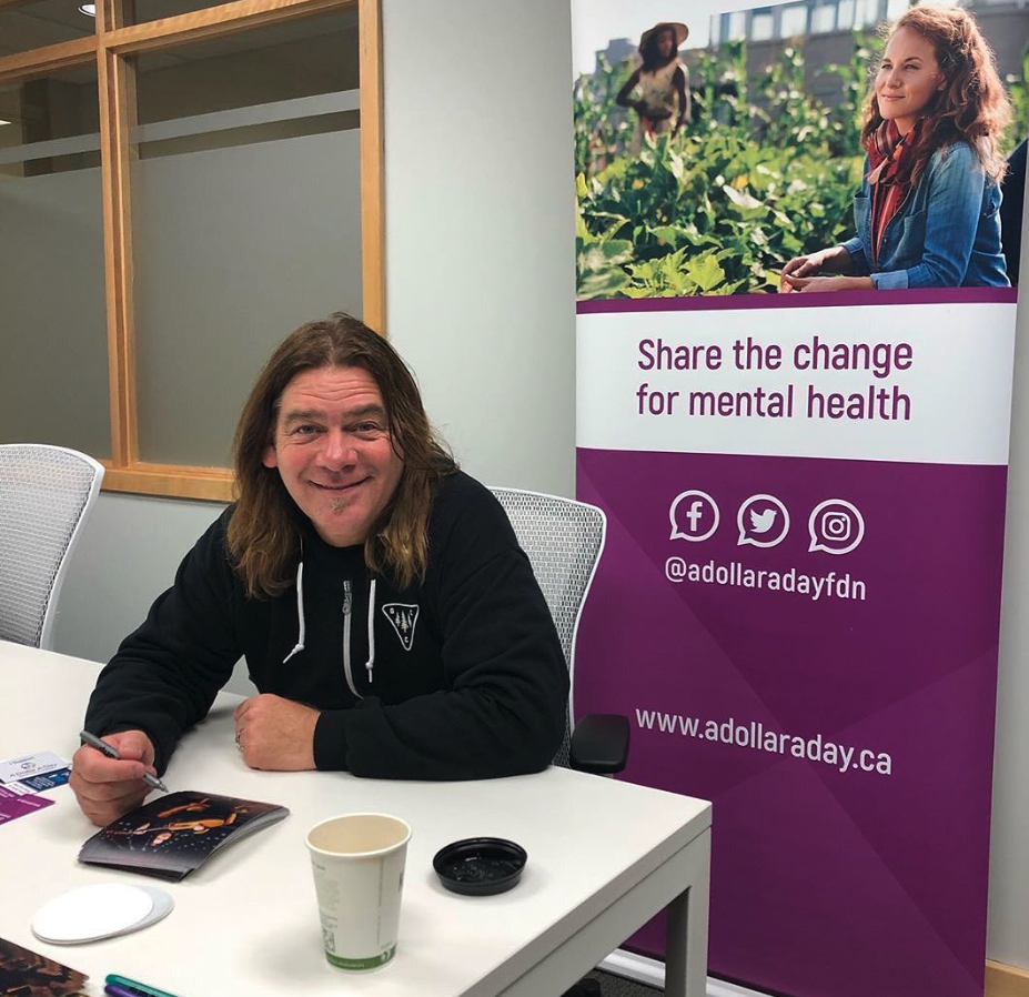A man with long brown hair, wearing a black hoodie, sitting at a white table with a pen, a paper, a disposable coffee cup, and a small black dish in front of him. Behind him is a purple banner promoting mental health awareness with social media icons and a website link. The corner of a white office chair is visible to his right, and a window with frosted glass panes is behind him.