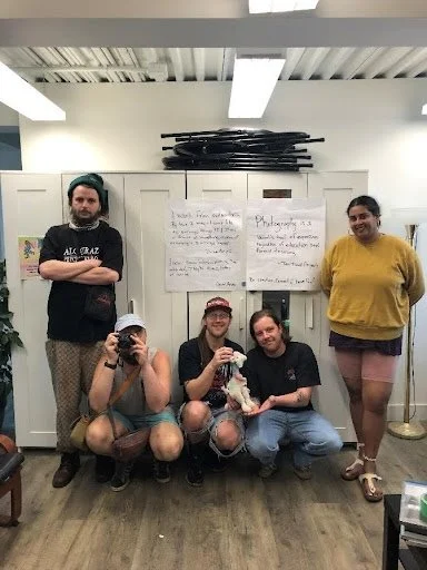 Group of five young people in a room with white lockers and posters on the wall, some sitting and some standing, posing for the photo.