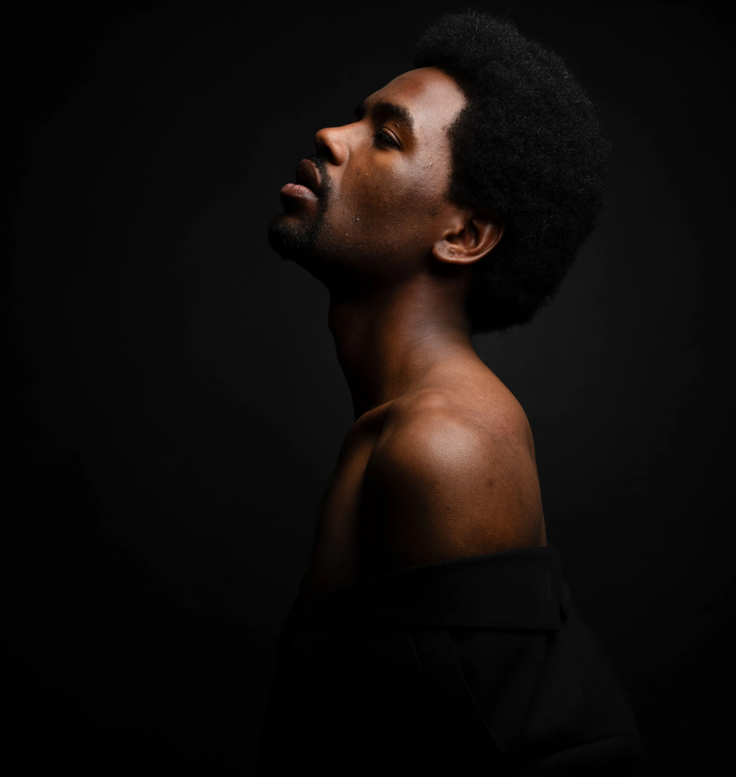 Side profile of a person with dark skin, natural curly hair, and a contemplative expression, against a black background.