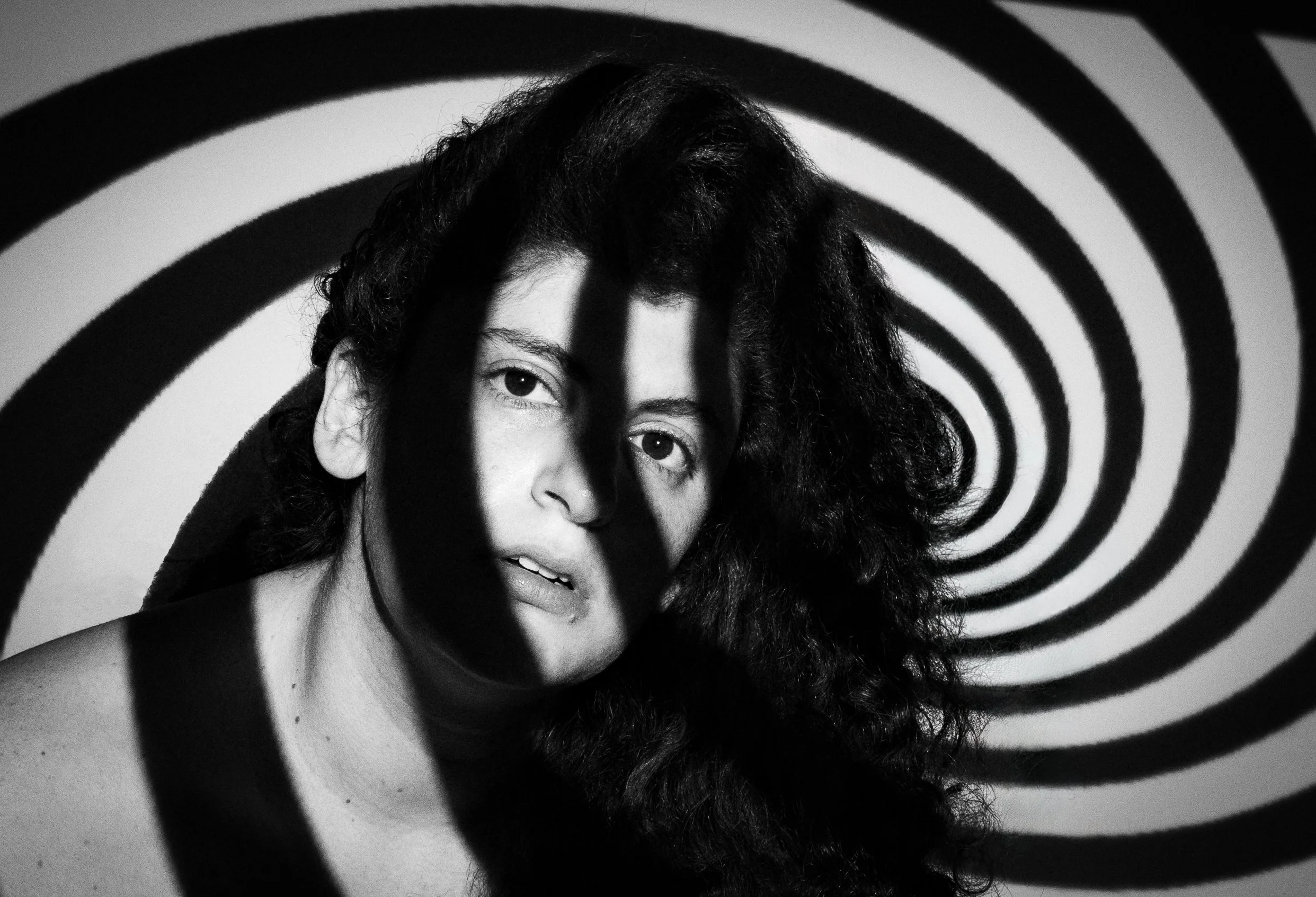 Black and white photo of a woman with curly hair behind a spiral pattern, partially obscuring her face.