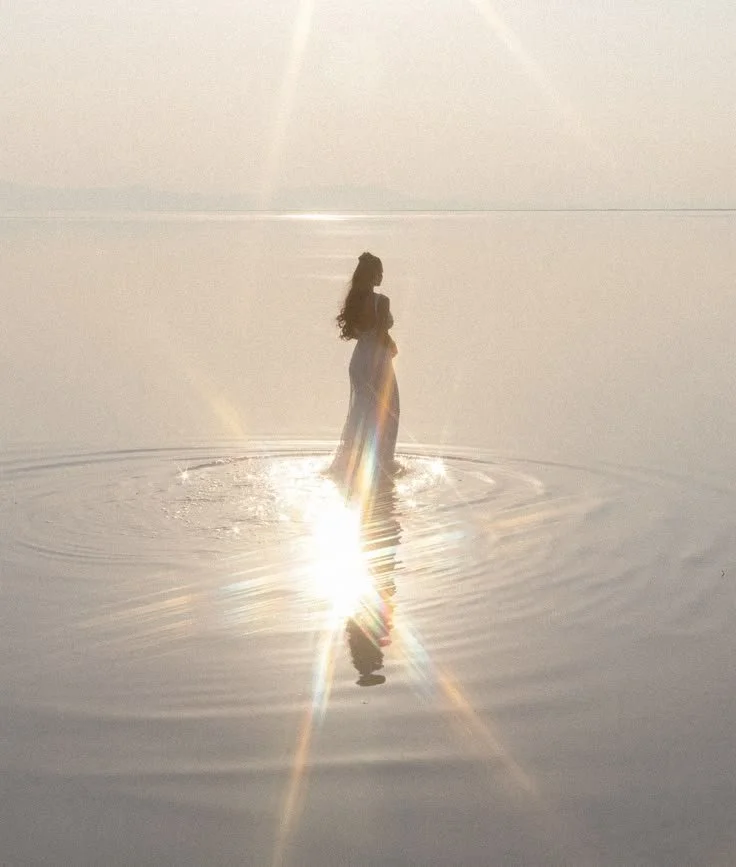 A woman standing in shallow water at sunset or sunrise, creating a silhouette with the sun reflecting on the water, producing starburst effects.