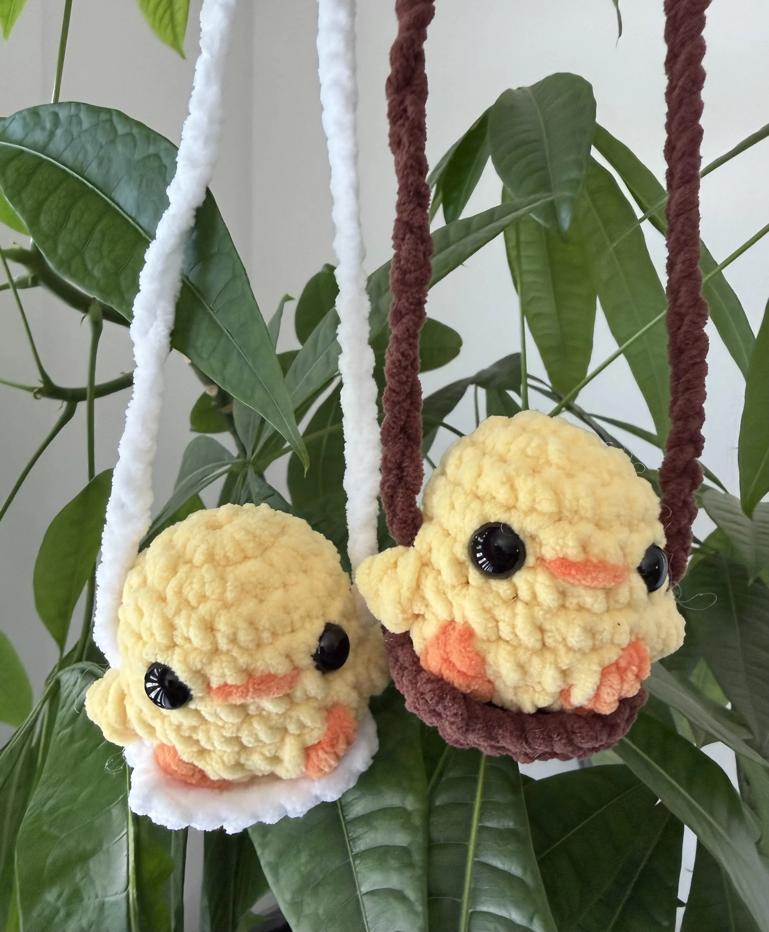Decorative hanging plush duck toys with large black eyes, orange beak, and fuzzy textured body, surrounded by large green leaves.