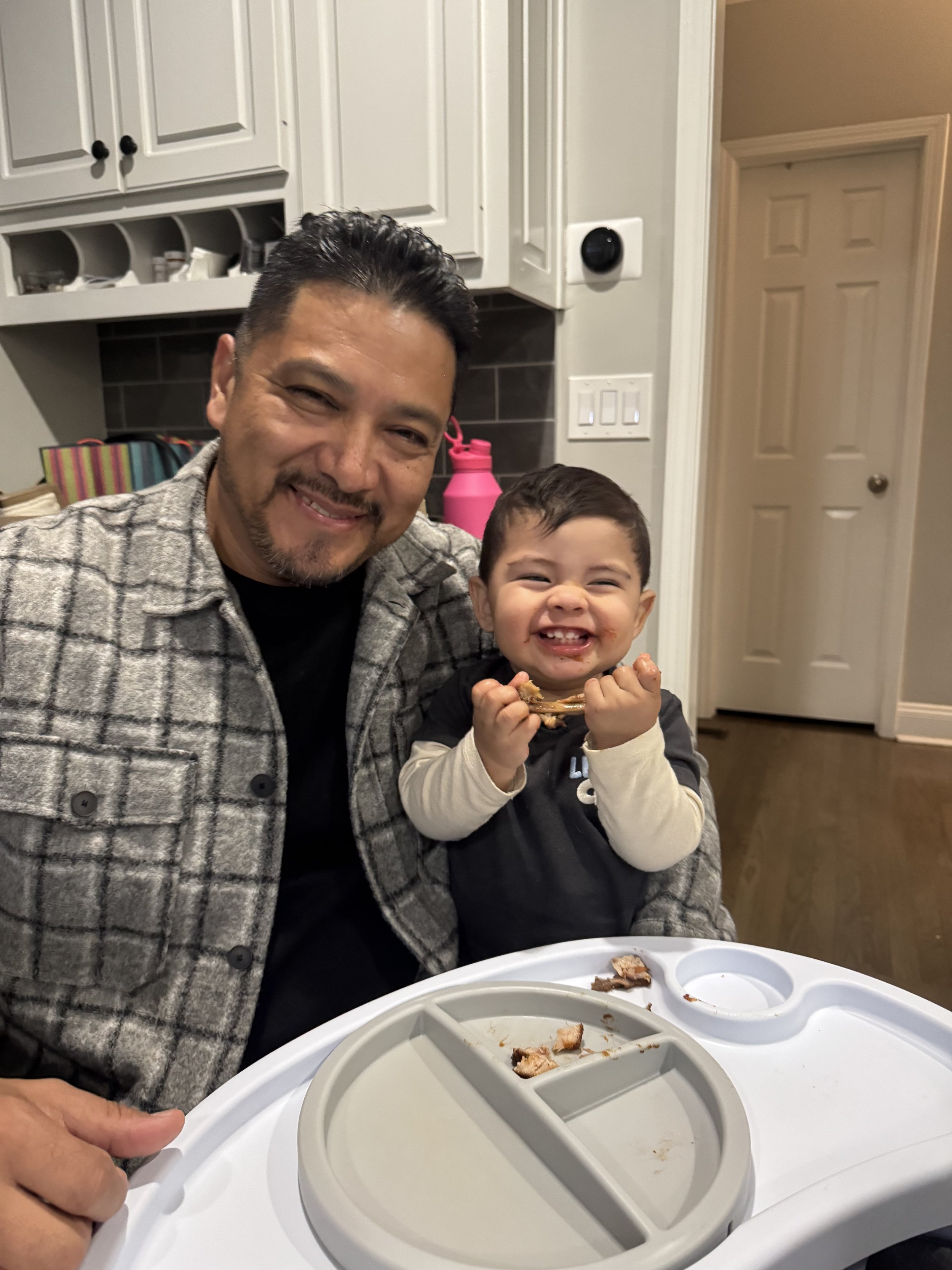 A smiling man and young boy enjoying a meal together in a kitchen, with an ice cream maker on the table and a door in the background.