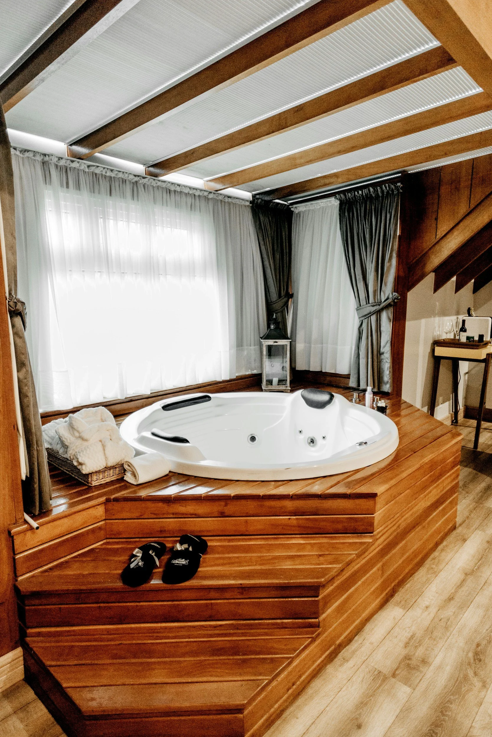 A wooden platform with a white hot tub, a basket of towels, and slippers near a large window with white curtains.