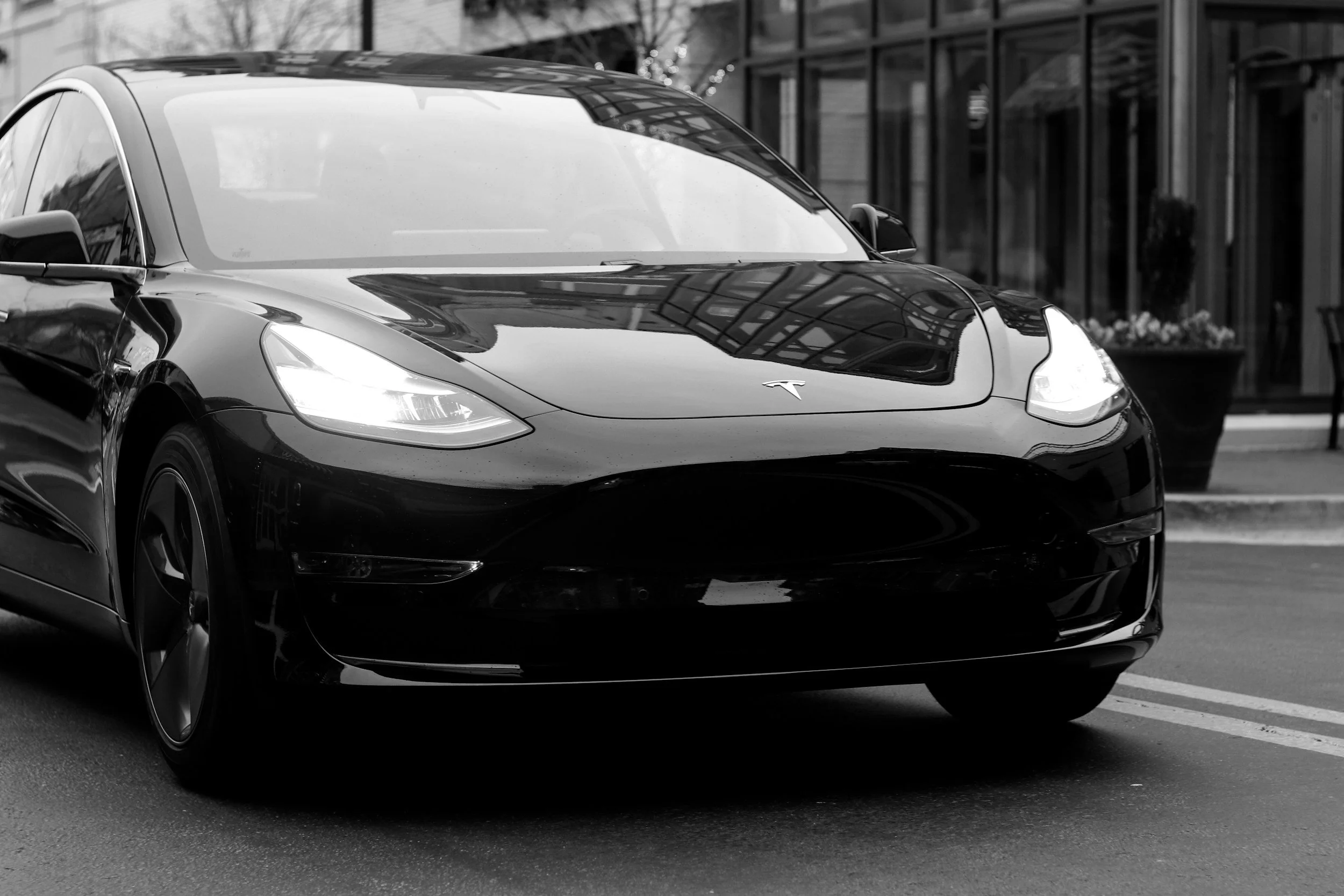 A black Tesla car parked outside a building with glass windows, reflection of the building visible on the car's surface, black and white photo.