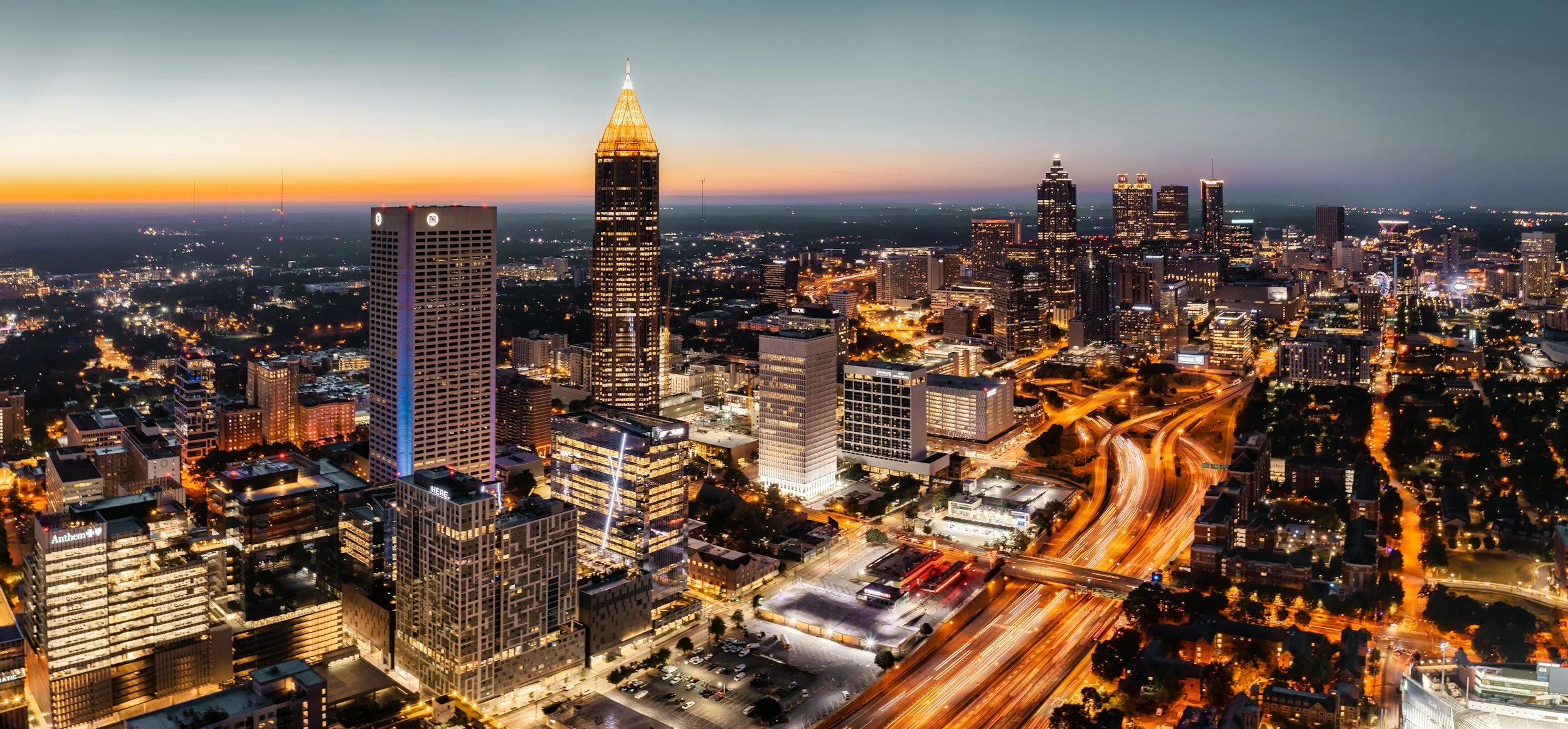 Nighttime aerial view of a city skyline with illuminated skyscrapers, busy roads with light trails, and a sunset sky in the background.