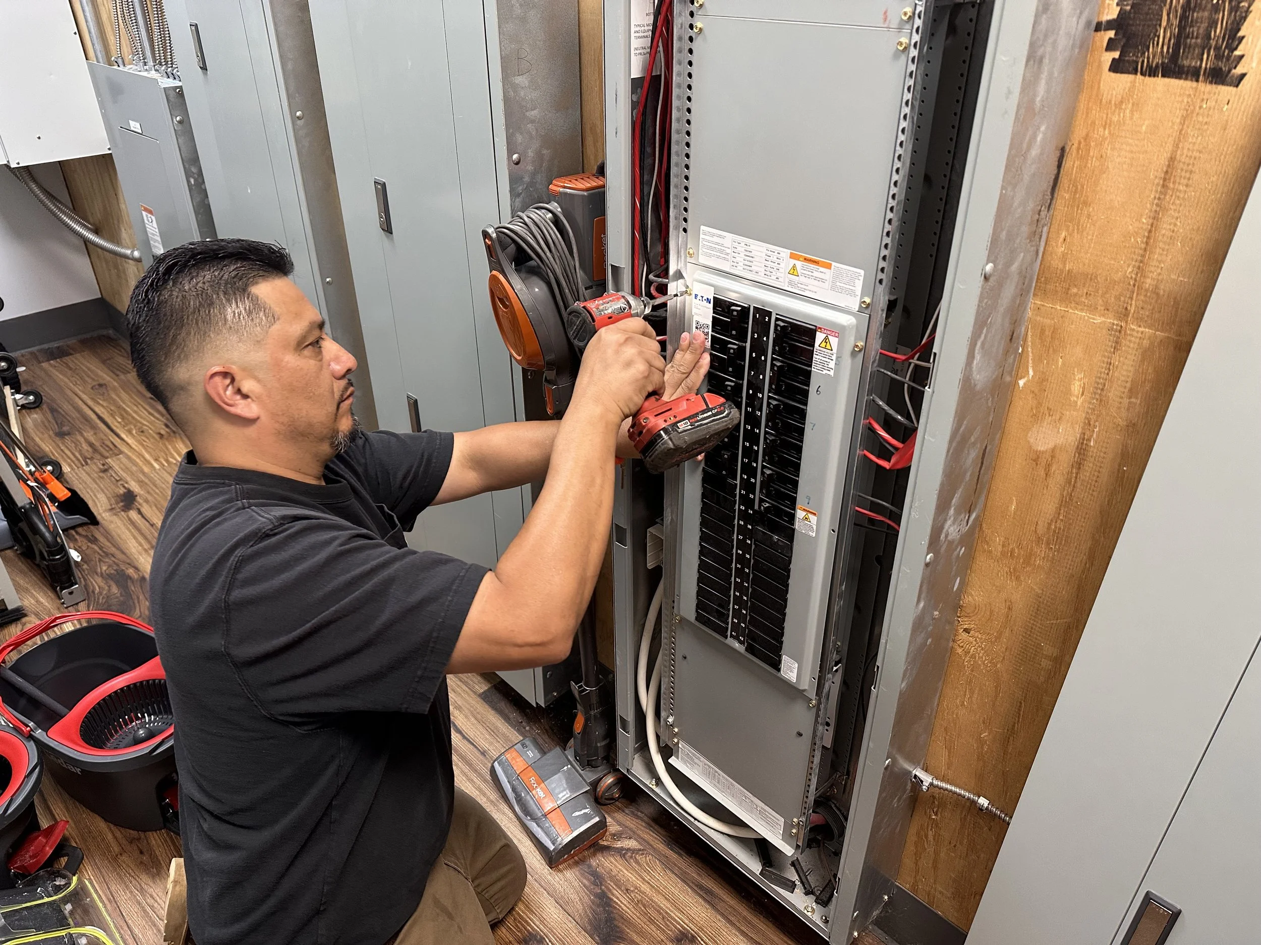 A technician working on an electrical panel with a cordless drill in hand. The panel contains multiple circuit breakers and wiring. Tools and equipment are nearby on the wooden floor.