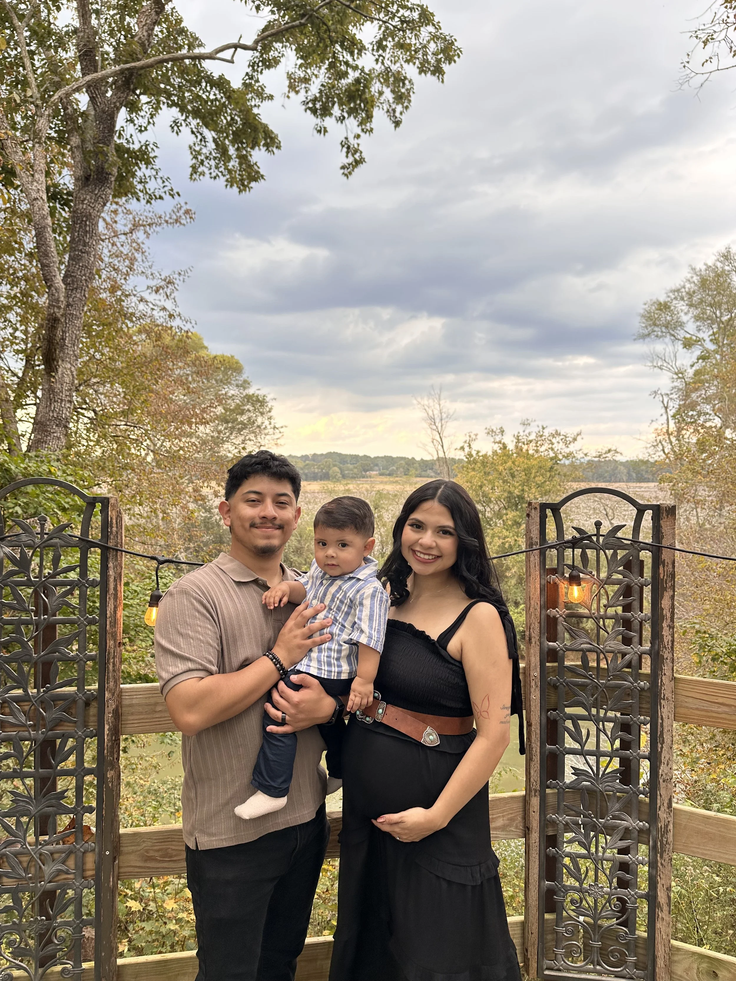 A family of three stands outdoors on a wooden bridge with decorative iron gates. The man is holding a young boy, and a woman stands beside them. They are smiling, with trees and a cloudy sky in the background.