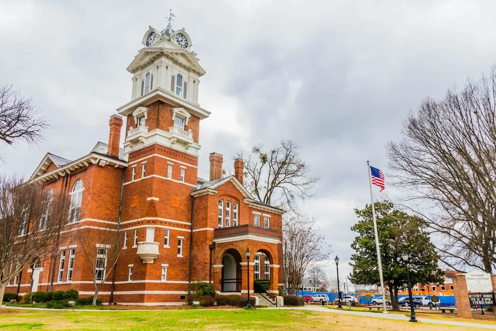 A historic red brick building with a tall clock tower, located on a cloudy day. There are leafless trees and an American flag on a flagpole nearby.