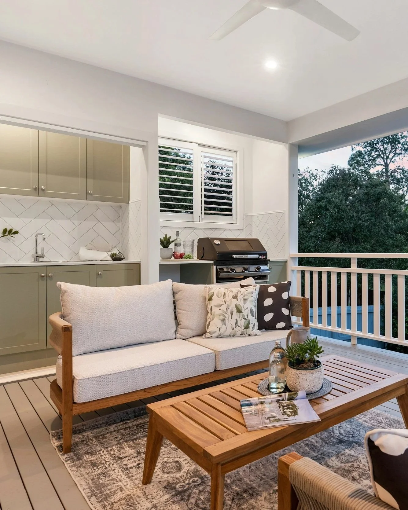 A cozy outdoor patio with a sofa, coffee table, and a grilling station with greenery visible outside.