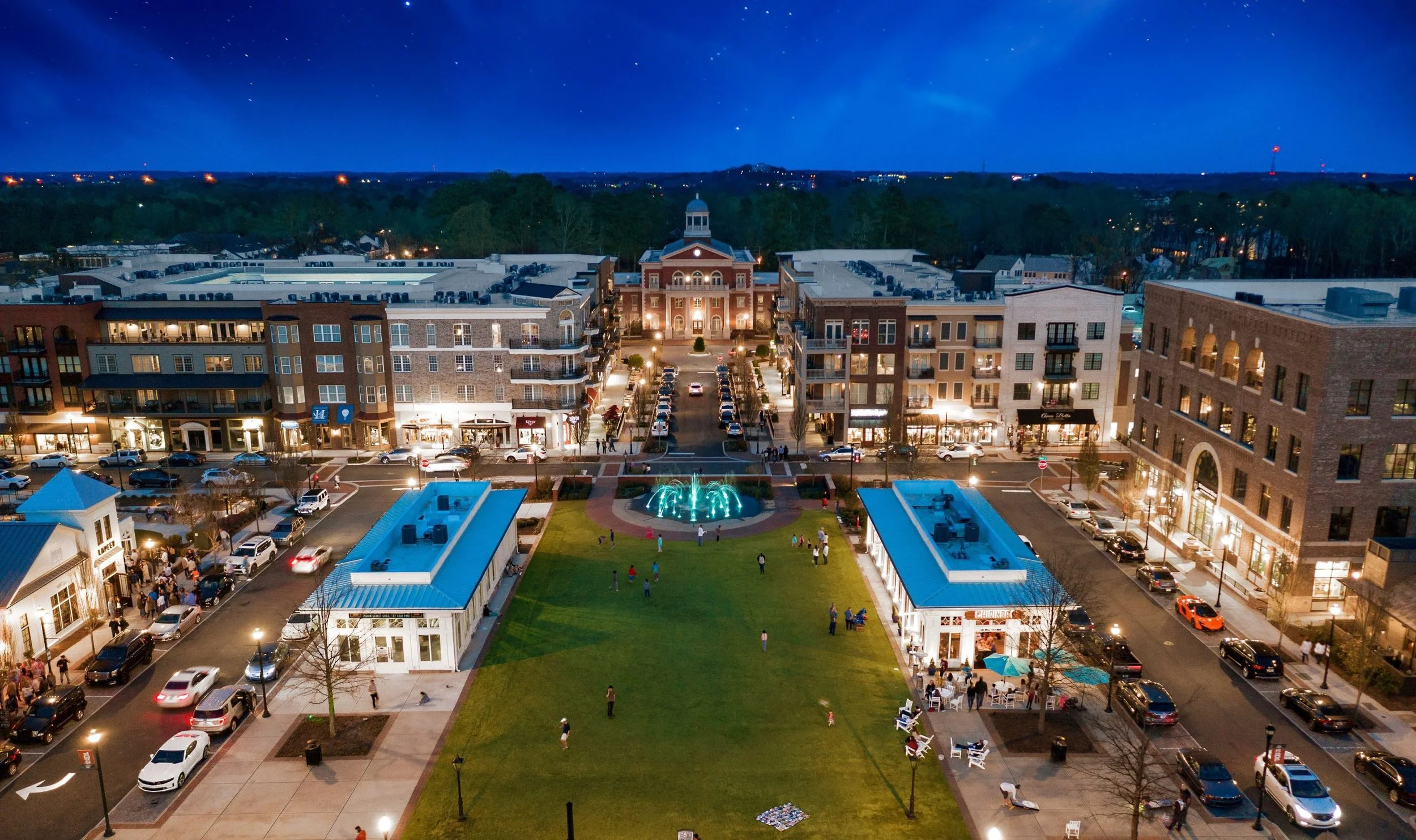 Night view of a city square with a green park and a water fountain, surrounded by shops, and multi-story buildings, with cars on the streets and a clear starry sky overhead.