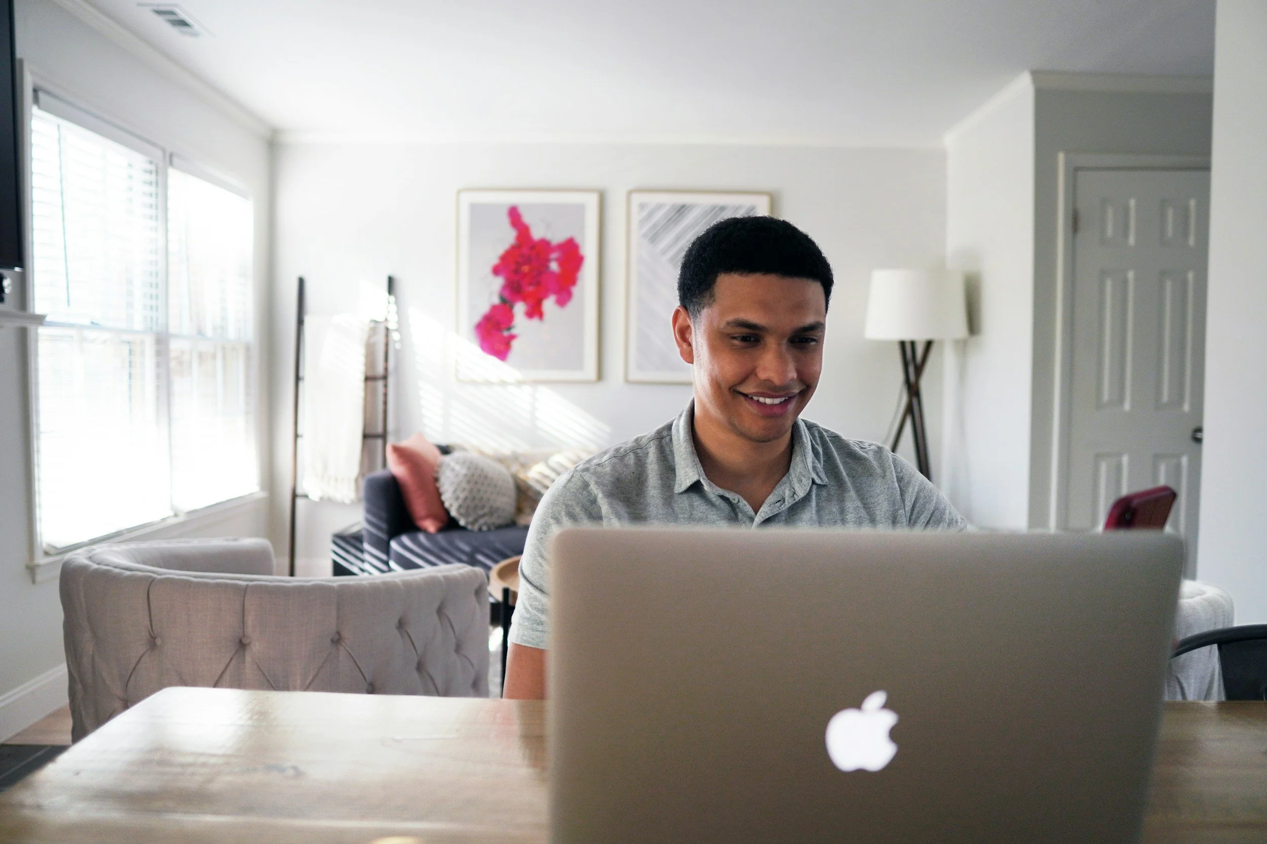 A young man sitting at a wooden table using a laptop in a bright, modern living room. There are large windows with blinds, a couch with cushions, framed artwork on the wall, and a standing lamp in the background.