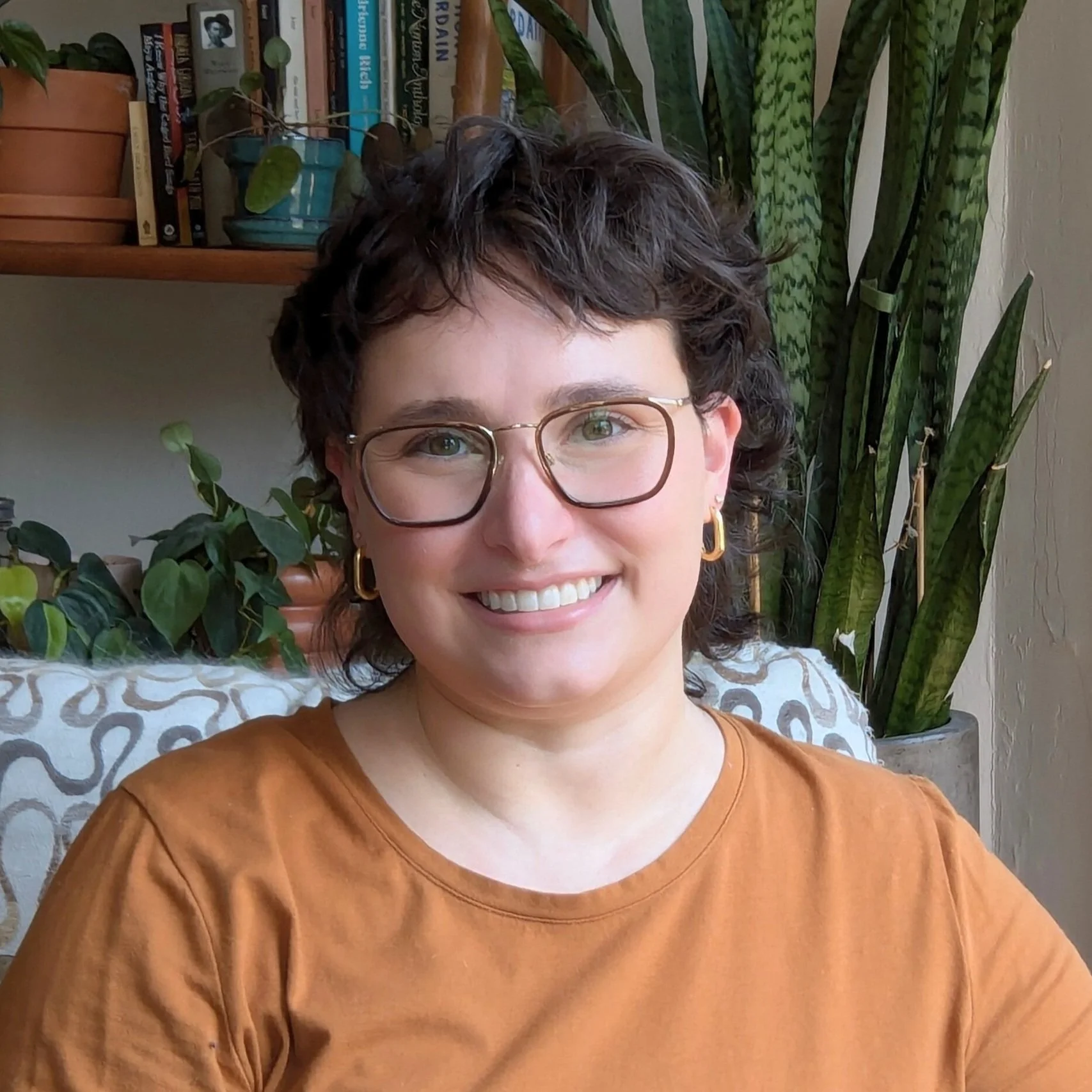 A woman with short curly dark hair, glasses, gold earrings, and a warm smile, sitting indoors with green houseplants and a bookshelf in the background.