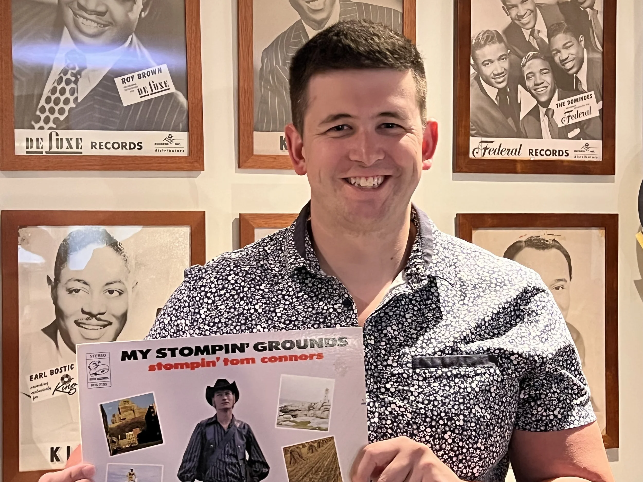 A smiling man holding a record album titled "My Stompin' Grounds" with a picture of a man in a cowboy hat on the cover, standing in front of a wall displaying vintage black-and-white portraits of musicians and singers.