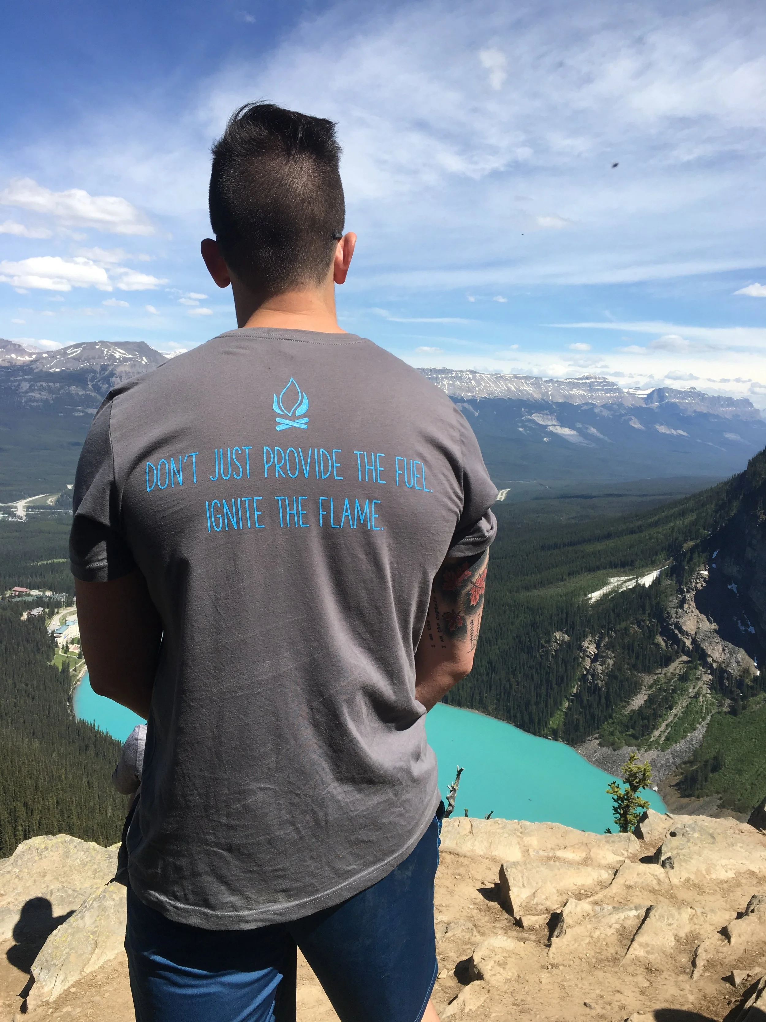 Ken standing on a cliff overlooking a green valley with a bright blue lake, snow-capped mountains in the distance, under a partly cloudy sky. They are wearing a gray t-shirt with blue text that reads 'Don't just provide the fuel. Ignite the flame.'