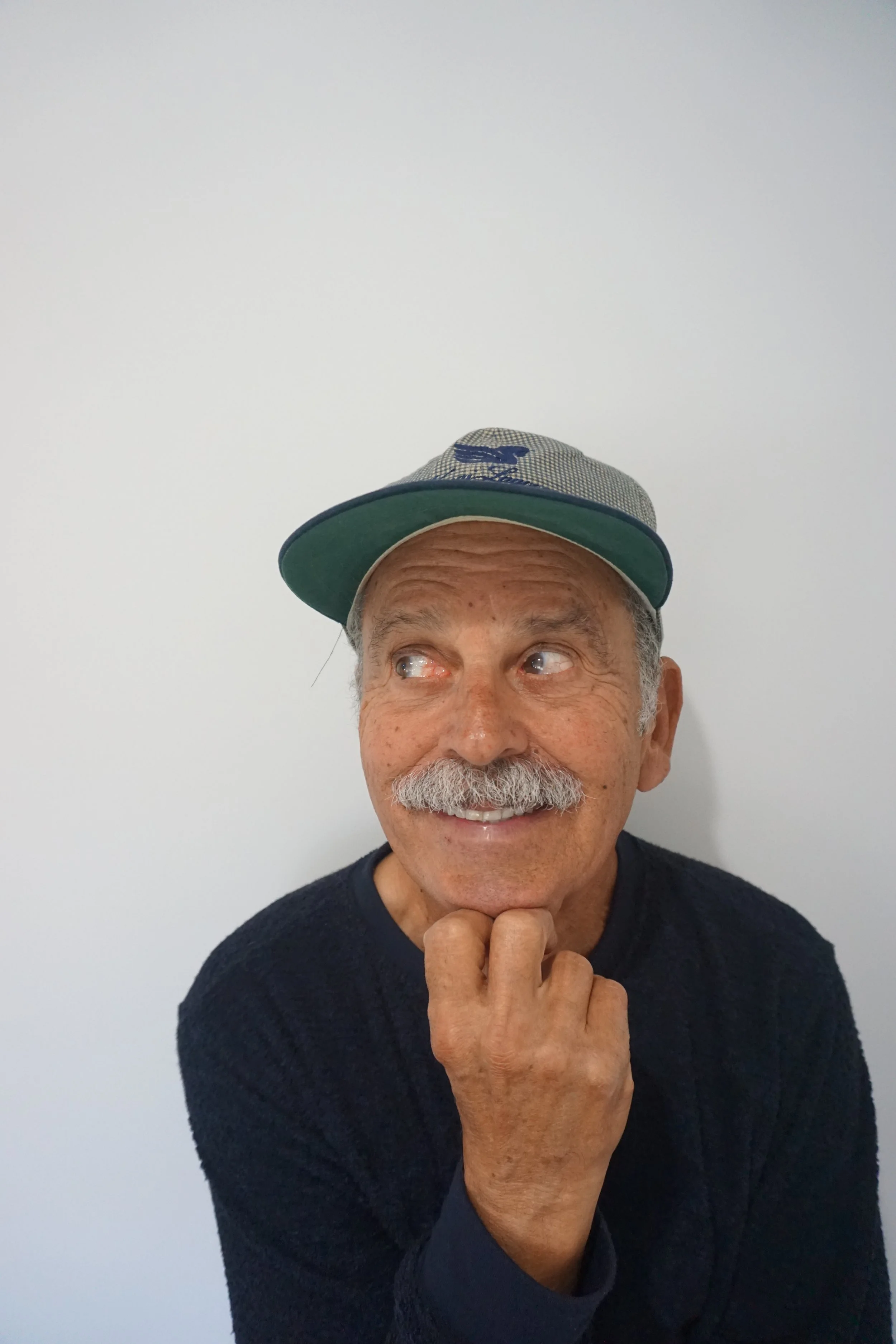 A smiling elderly man with a gray mustache and beard, wearing a gray and green baseball cap and a dark blue sweater, posing with his hand under his chin in front of a plain white wall.