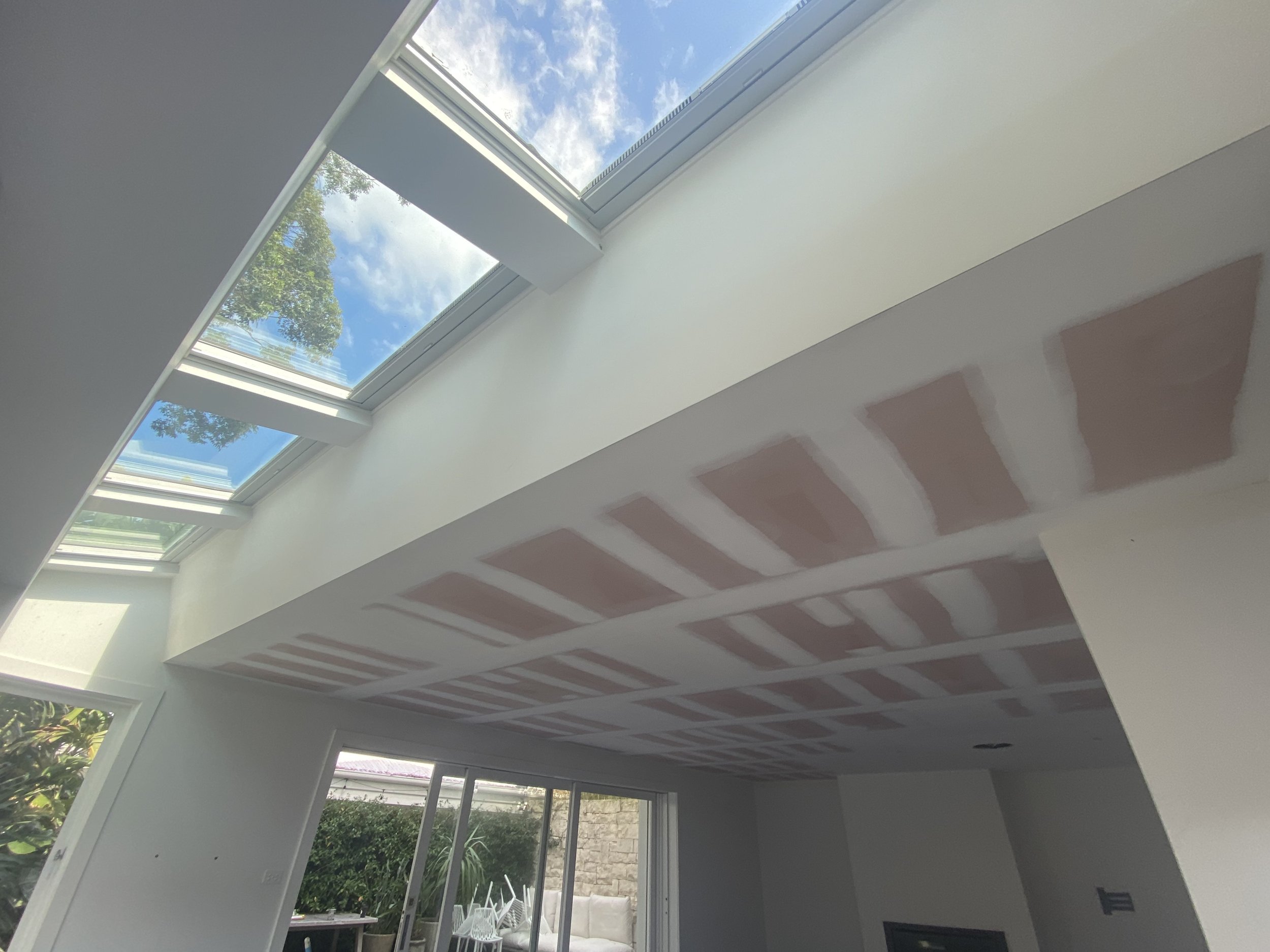 Interior of a room with a high sloped ceiling featuring a large skylight window, showing a partly cloudy sky and green tree outside. The ceiling has visible drywall joints and patches preparing for painting or finishing. The room includes a large window or sliding glass door at the back with a patio and garden outside.