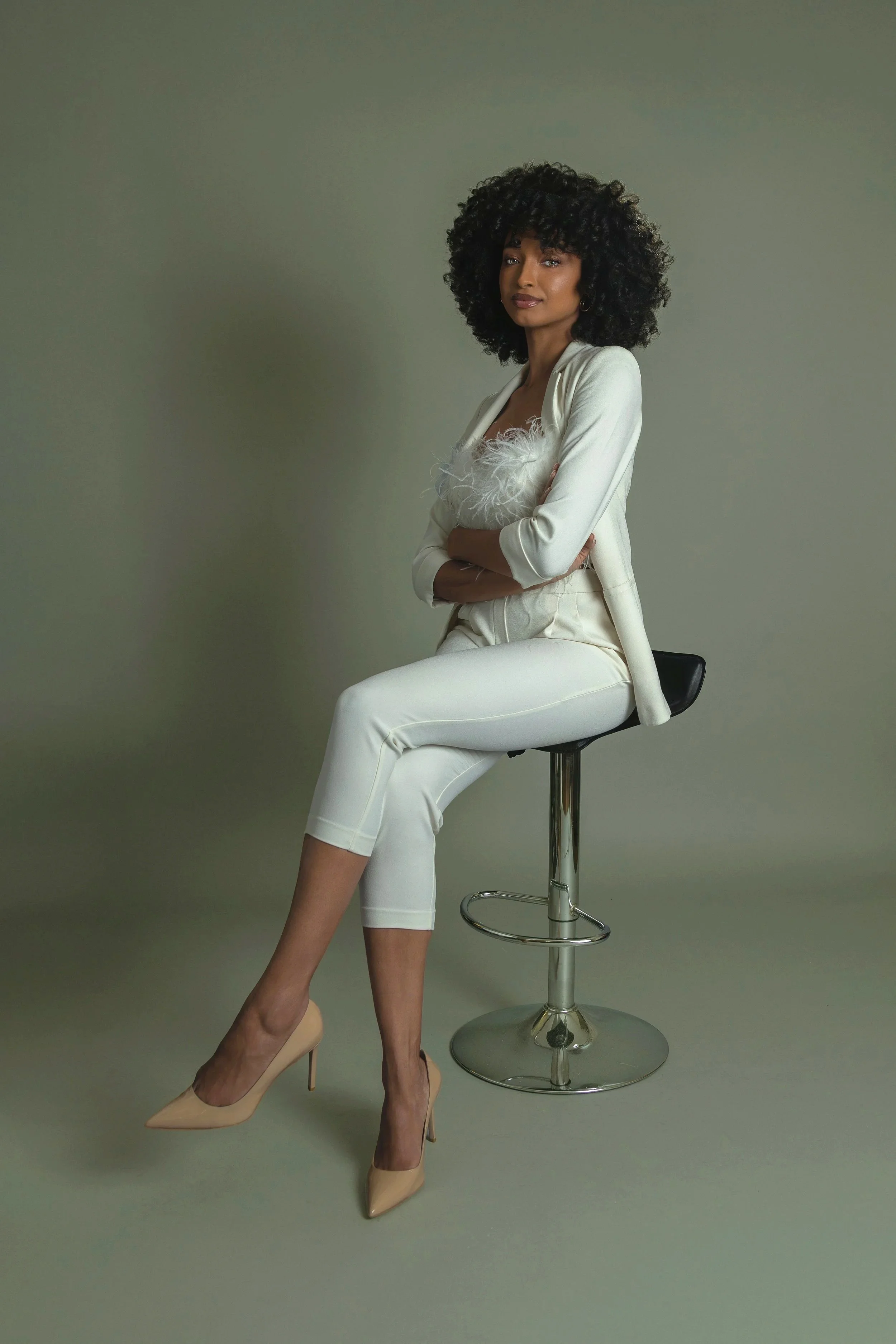 A woman with dark curly hair wearing a white blazer, white pants, and nude high heels sitting on a black stool against a gray background, holding a feathered white clutch.