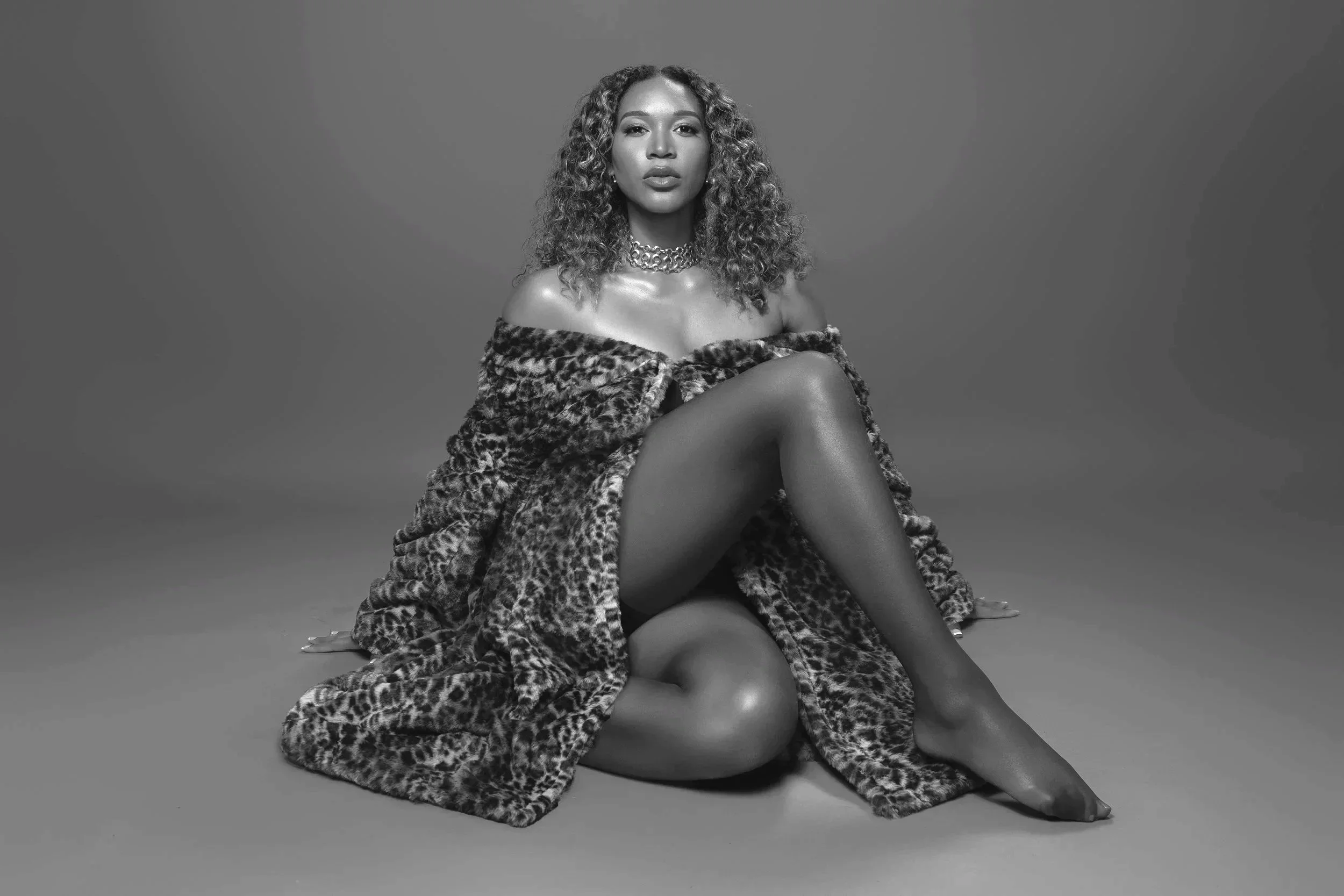 Black and white photo of a woman with curly hair wearing a leopard-print coat and a thick necklace, sitting against a plain background.