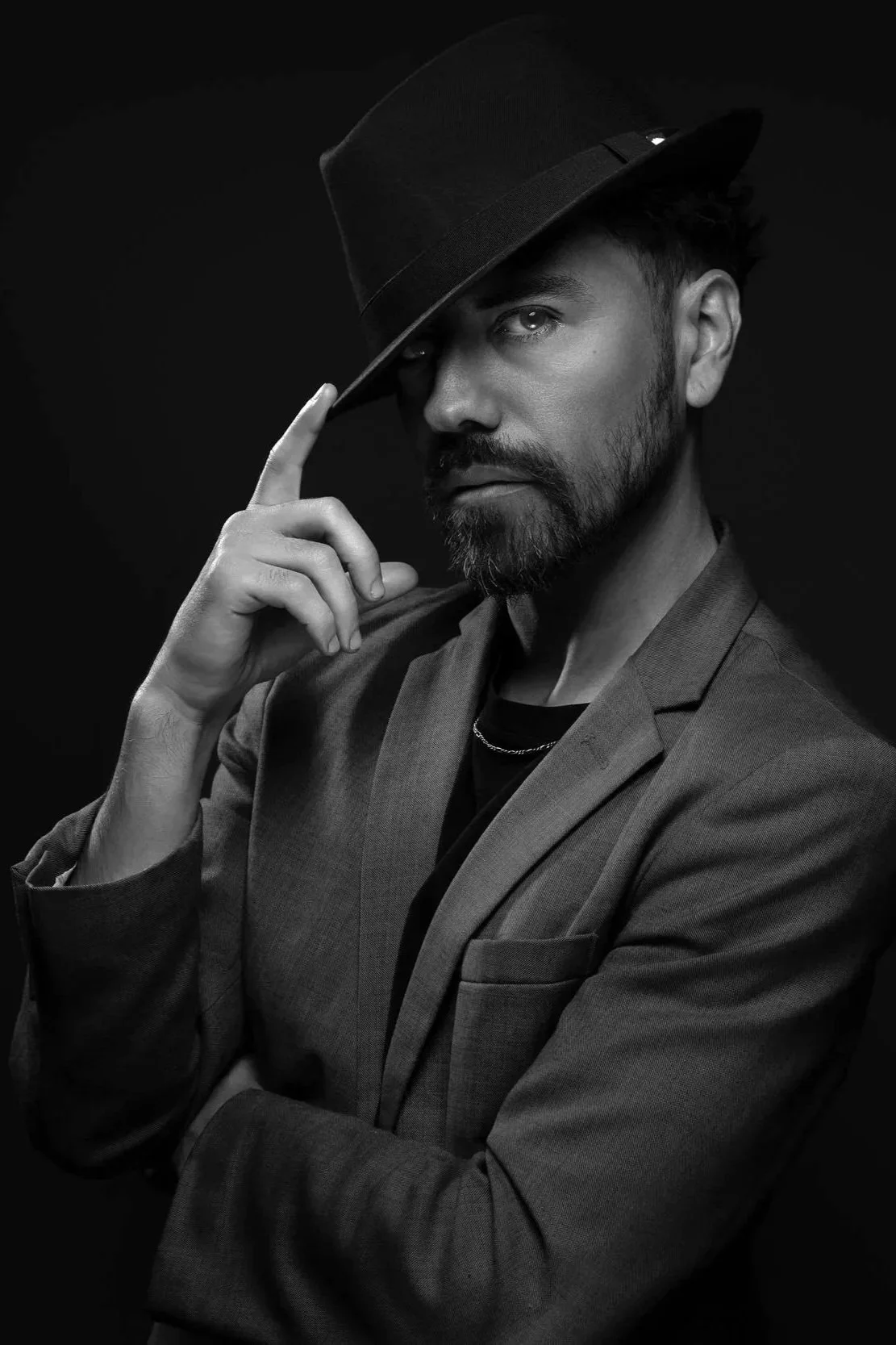 Black and white portrait of a man wearing a suit and fedora, touching the brim of his hat with one hand, with a serious expression, against a dark background.