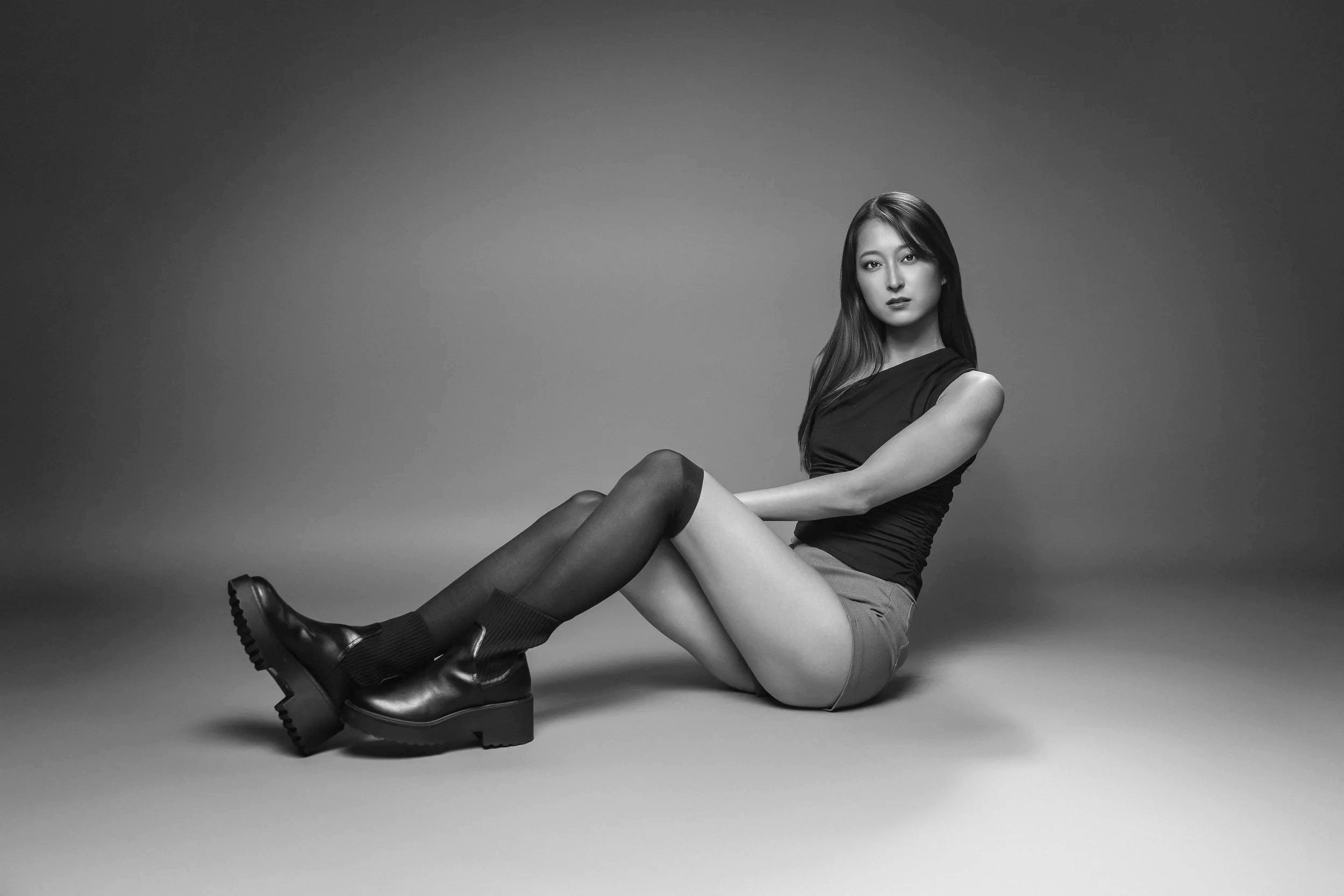 Black and white photo of a woman sitting on the floor with her knees bent, wearing a dark top, shorts, thigh-high stockings, and boots, against a plain gray background.