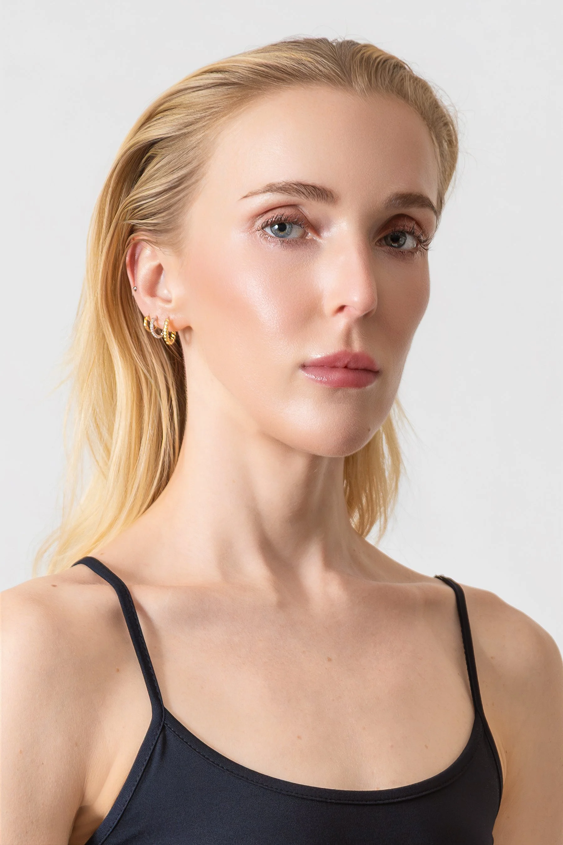 Close-up of a woman with blonde hair, blue eyes, wearing a black spaghetti strap top and earrings, against a plain white background.