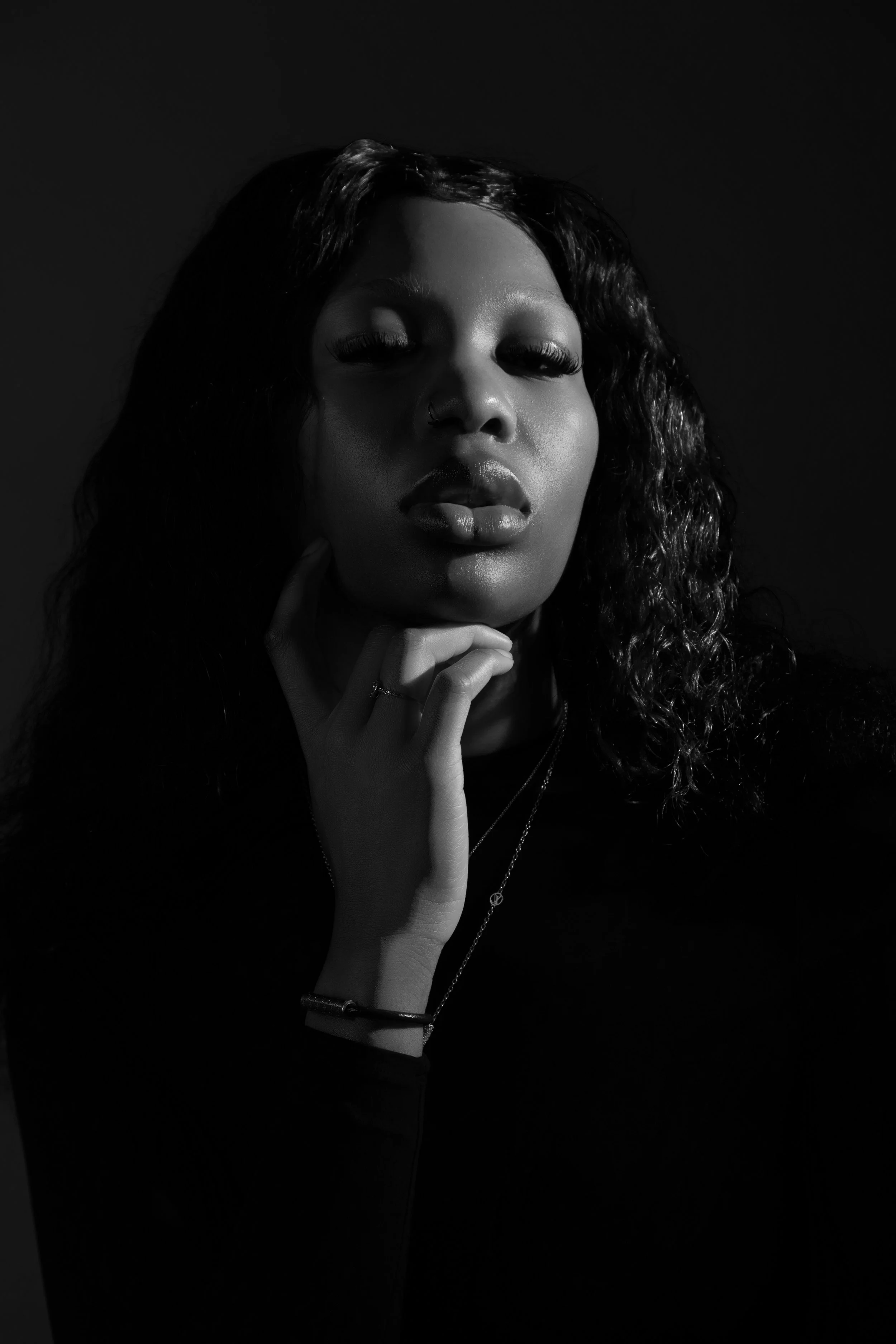 Black and white portrait of a woman with curly hair, wearing a nose ring, touching her chin with her right hand, against a dark background.
