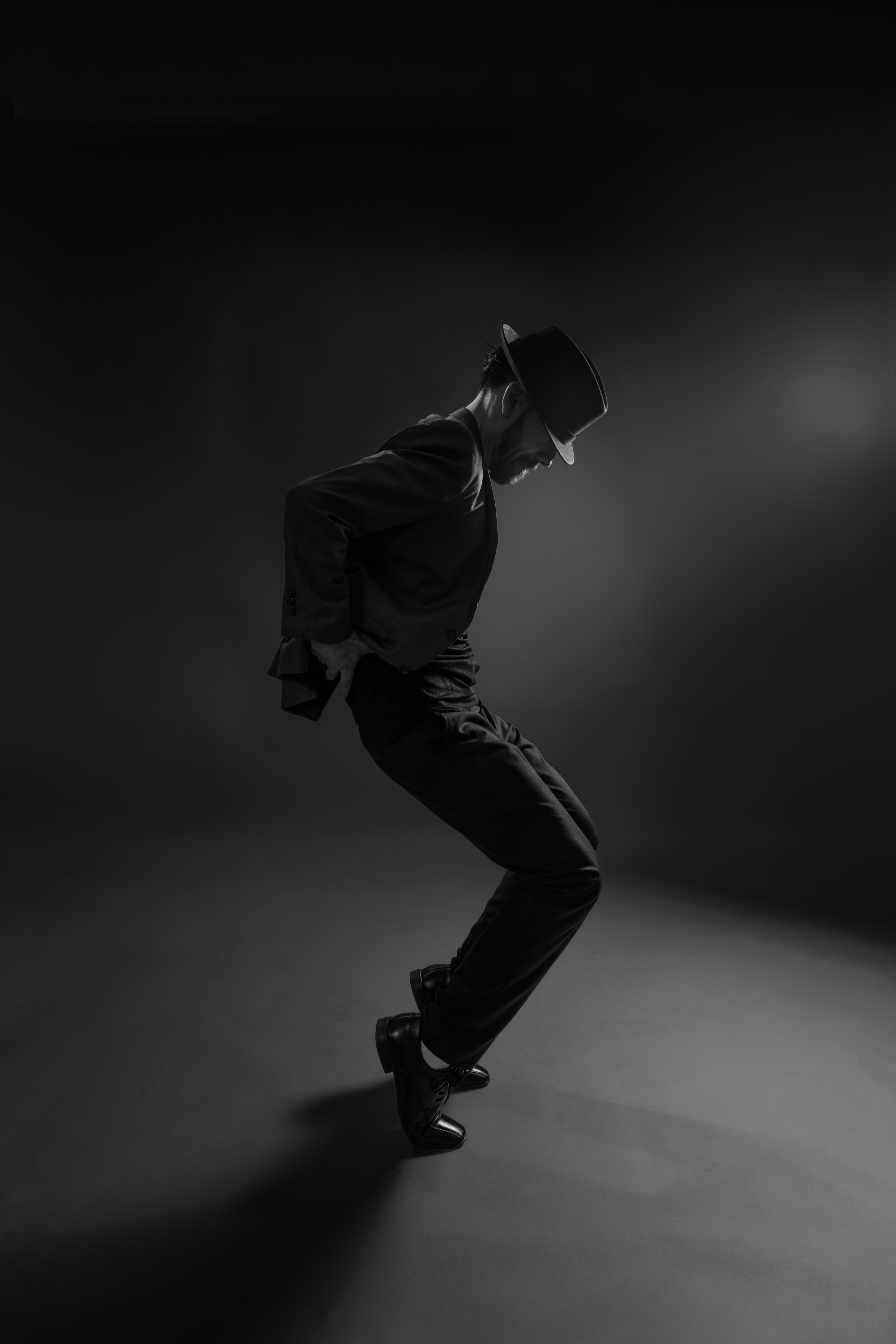 A black and white photo of a man dancing, wearing a suit, fedora, and boots, with his head tilted down and hands in his pockets, mid-movement.