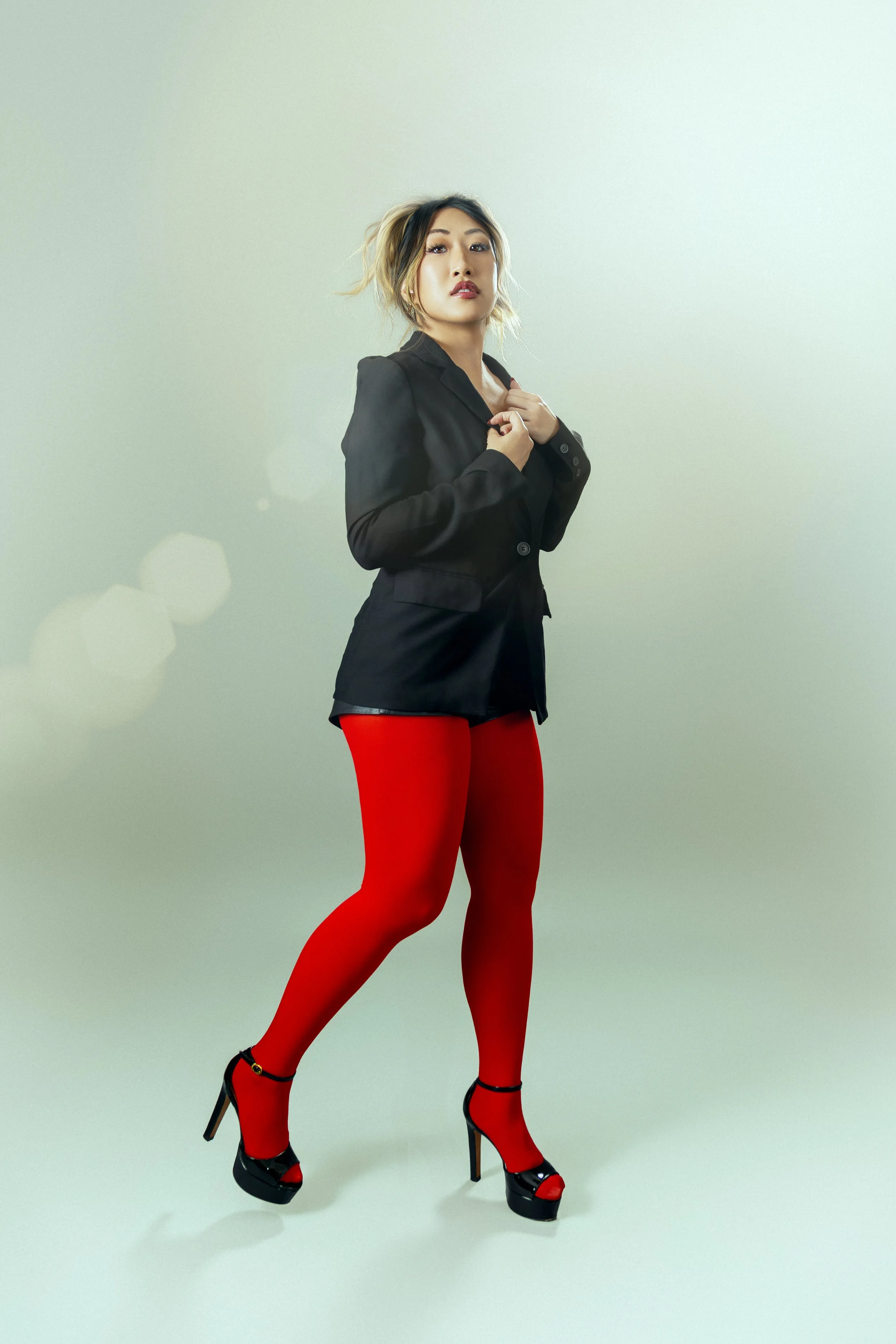 A woman wearing red tights, high-heeled shoes, a black blazer, and black lipstick, sets against a plain light green background.