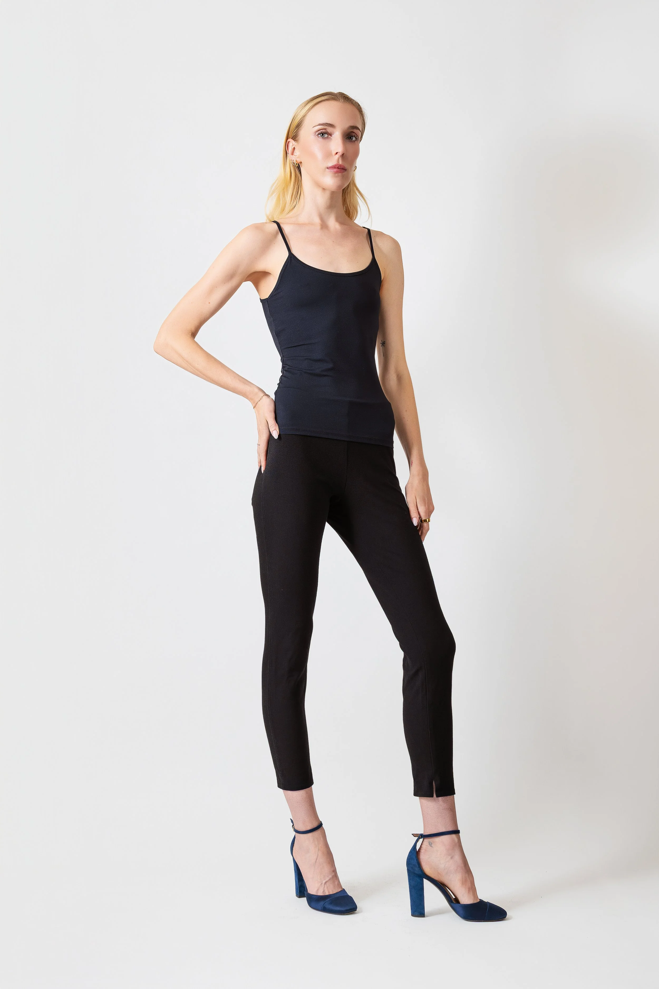 A woman with blonde hair wearing a black tank top, black pants, and blue high heels, posing against a plain white background.