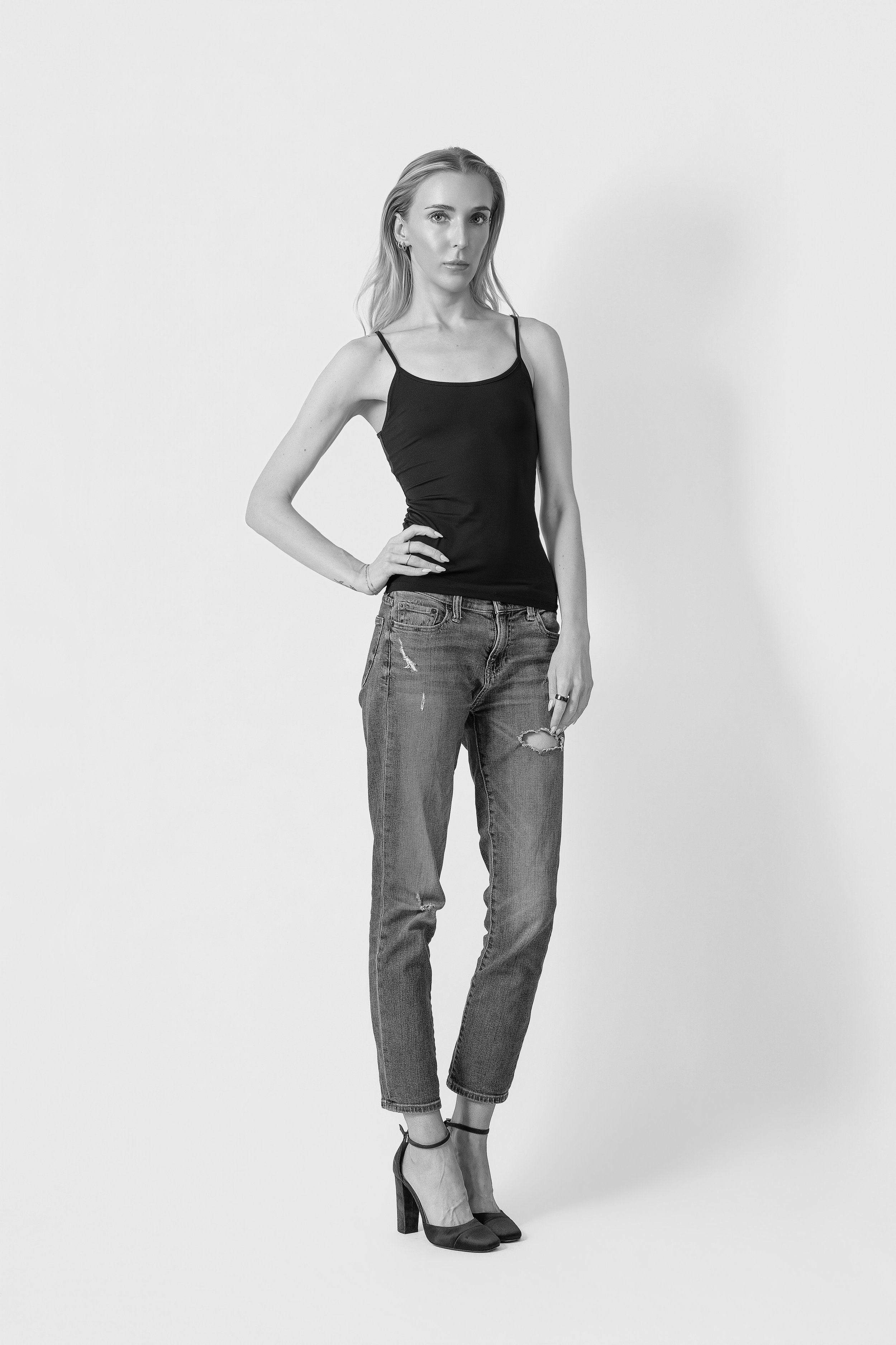 A woman with long hair wearing a black spaghetti strap top, ripped jeans, and high heels, standing against a plain white background.