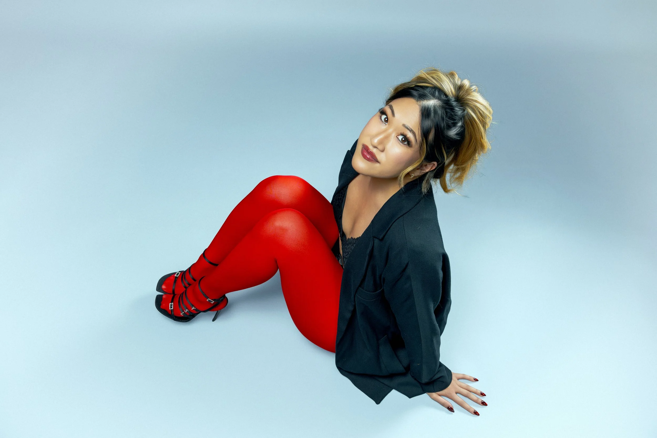 Portrait of a woman with shoulder-length hair, wearing red tights, black high-heeled shoes, and a black blazer, sitting on the floor against a light blue background.