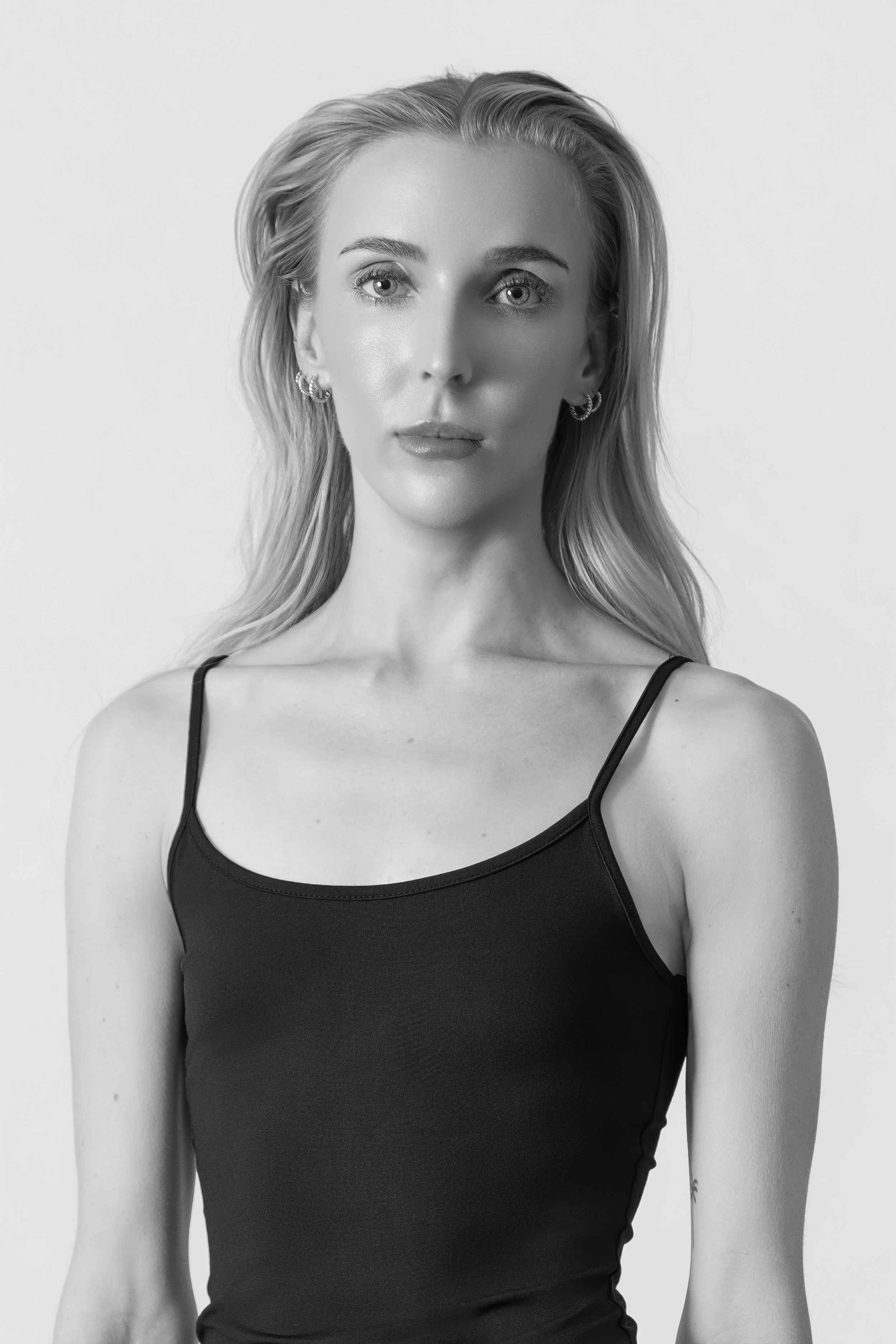 Black and white portrait of a woman with light-colored hair, wearing a black tank top and multiple earrings, looking directly at the camera.
