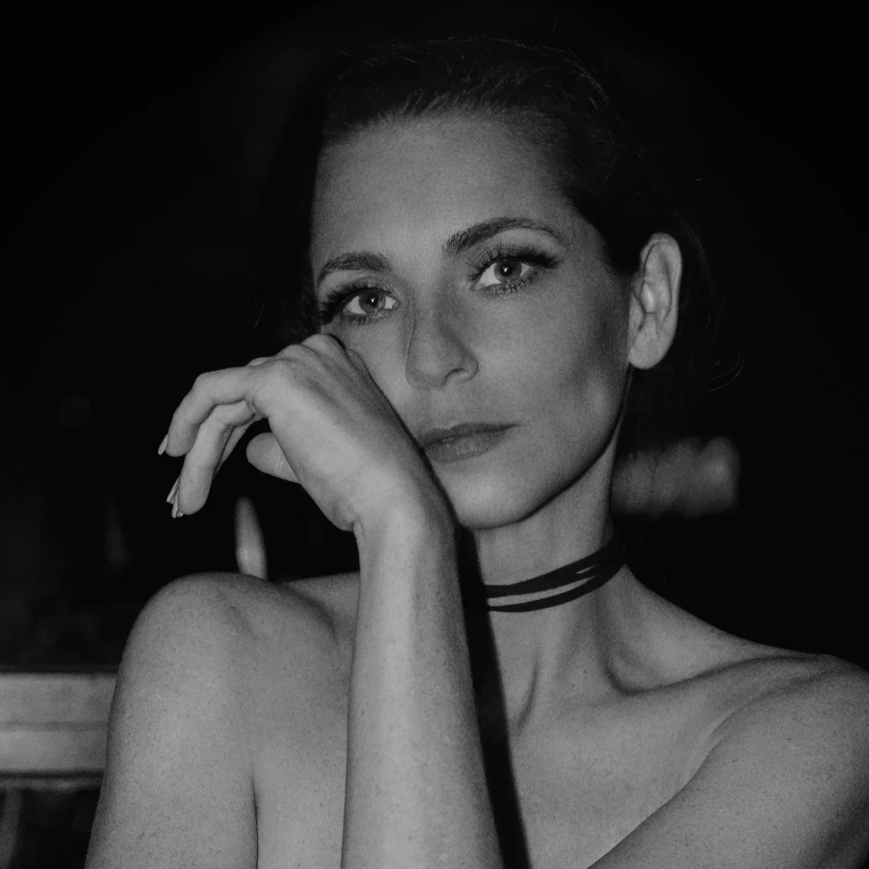 A black and white portrait of a woman with dark hair, wearing eyeliner and a striped choker, resting her face on her hand with intense eyes.