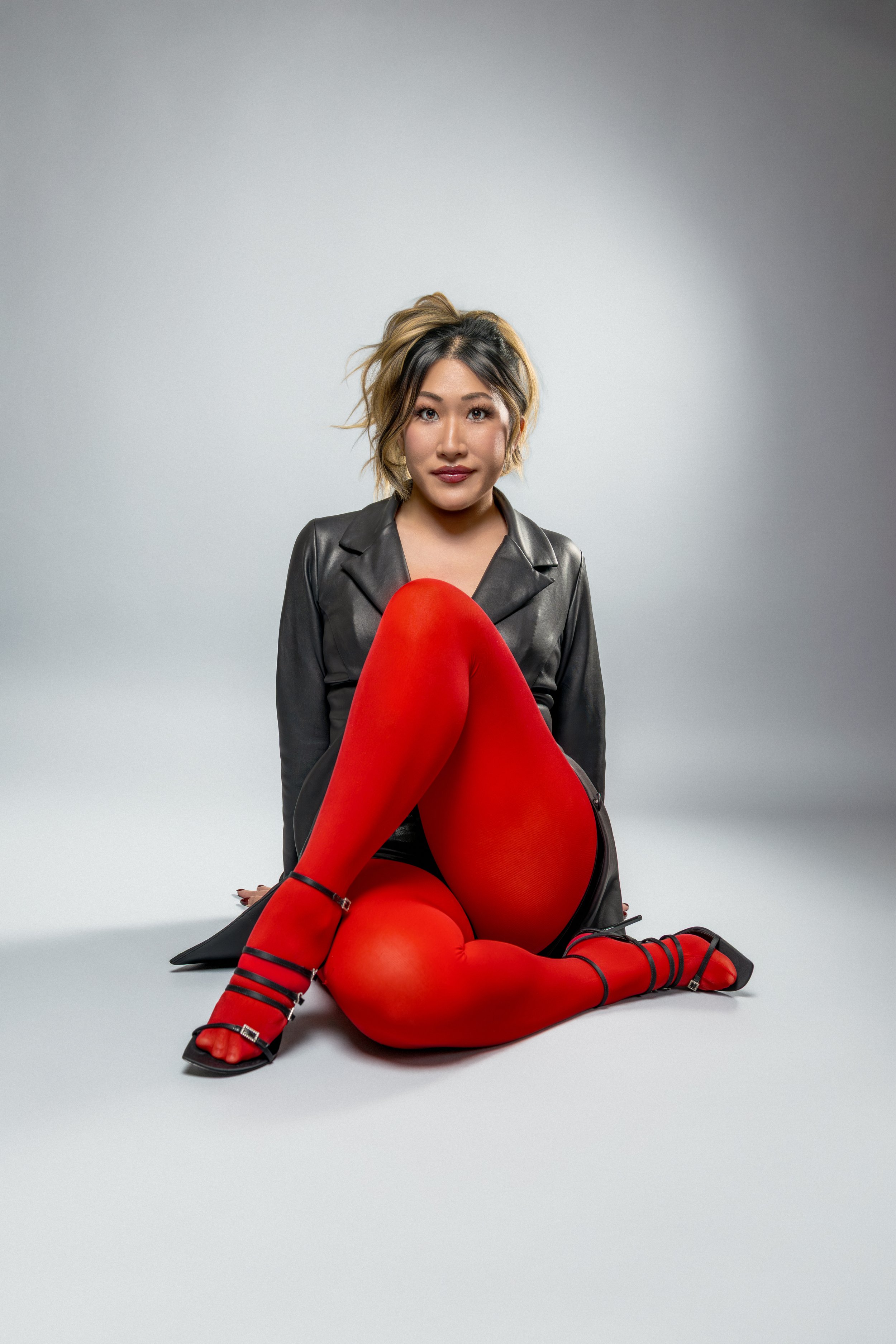 A woman with shoulder-length hair sitting on the floor in front of a plain gray background. She is dressed in a black leather jacket, red thigh-high stockings, and black strappy high heels.