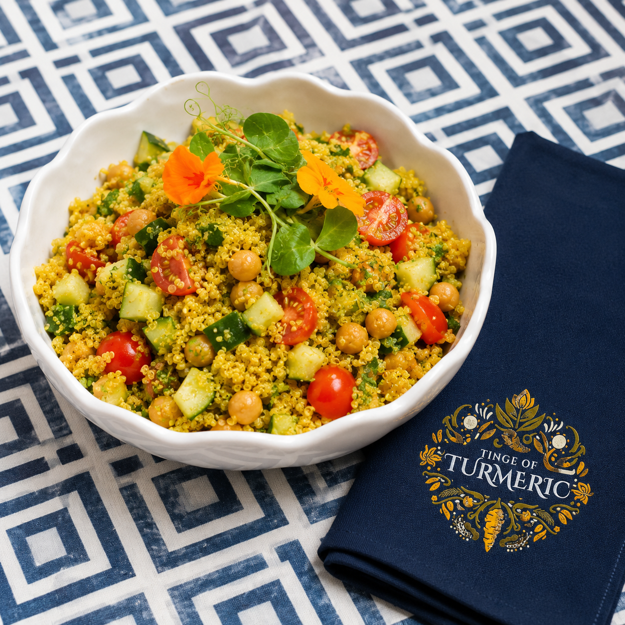 A bowl of tabbouleh salad with cherry tomatoes, cucumber, chickpeas, and fresh herbs, garnished with edible flowers and microgreens, placed on a table with a blue and white patterned tablecloth and a navy napkin with a turmeric label.