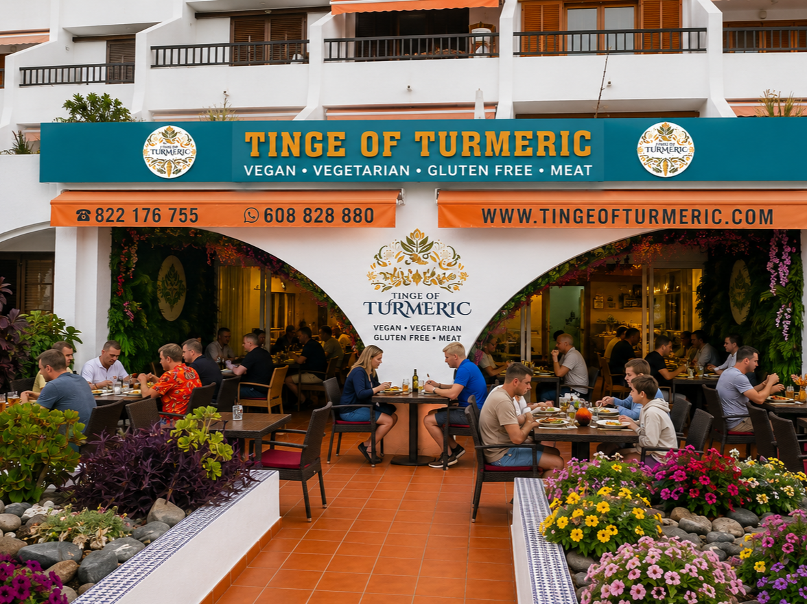 Outdoor dining area of a restaurant called Tinge of Turmeric, with people eating at tables under green and purple plants and colorful flowers.